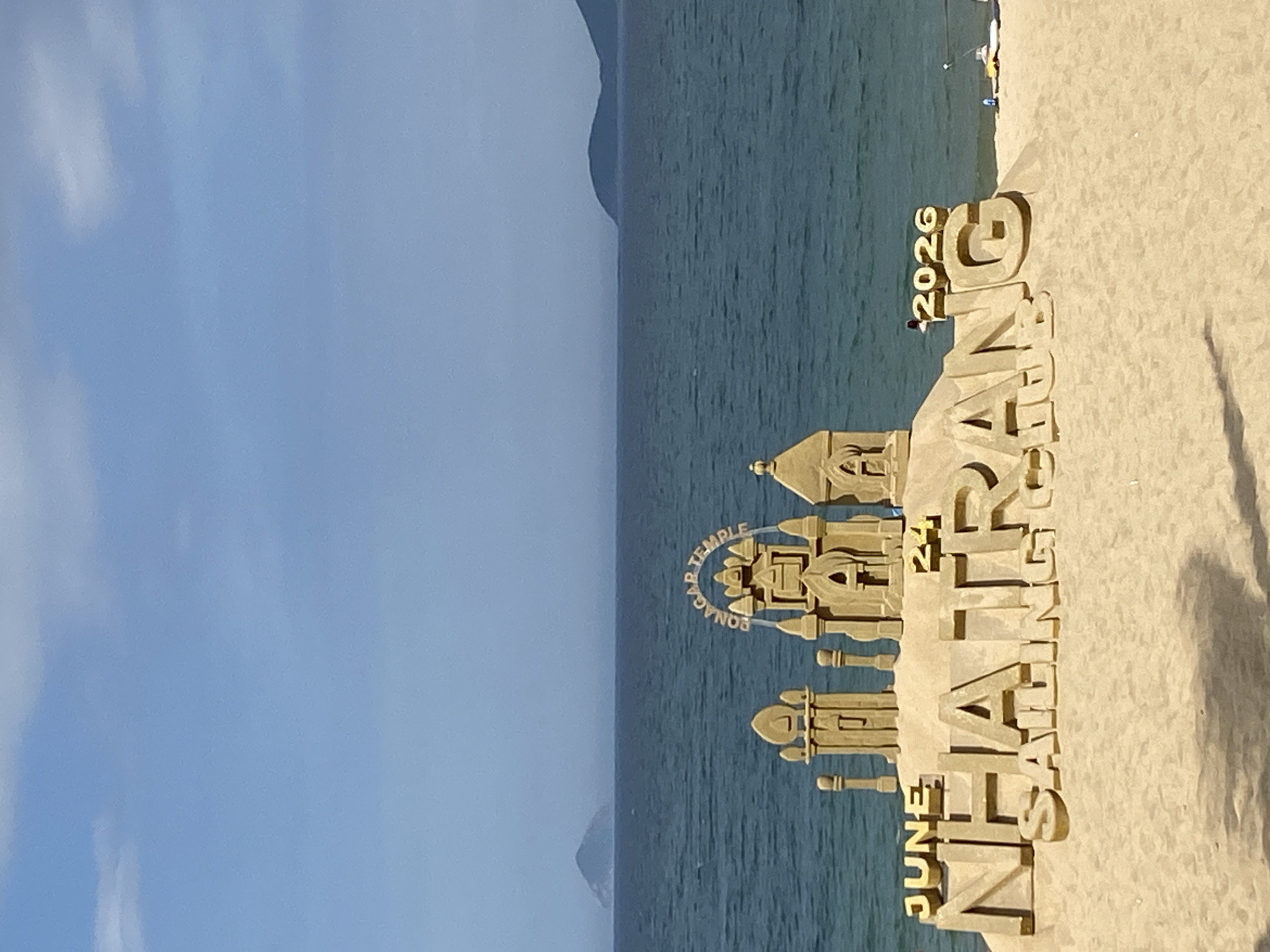 This image shows a sand sculpture on a beach in Nha Trang, Vietnam. The sculpture depicts a stylized temple and large lettering. Visible text includes "JUNE", "NHA TRANG", "2026", and "KONARK TEMPLE". The sculpture is made of sand, suggesting it is a temporary installation. The background shows the ocean with gentle waves and a clear, blue sky. The lighting suggests it is daytime, likely sunny, given the bright sand. The overall mood is peaceful and scenic, highlighting a tourist attraction or an event related to the year 2026 and the Konark Temple. There are a few small figures of people near the sculpture and in the distance on the beach, adding a sense of scale. The environment is a typical beach setting with sand and water.