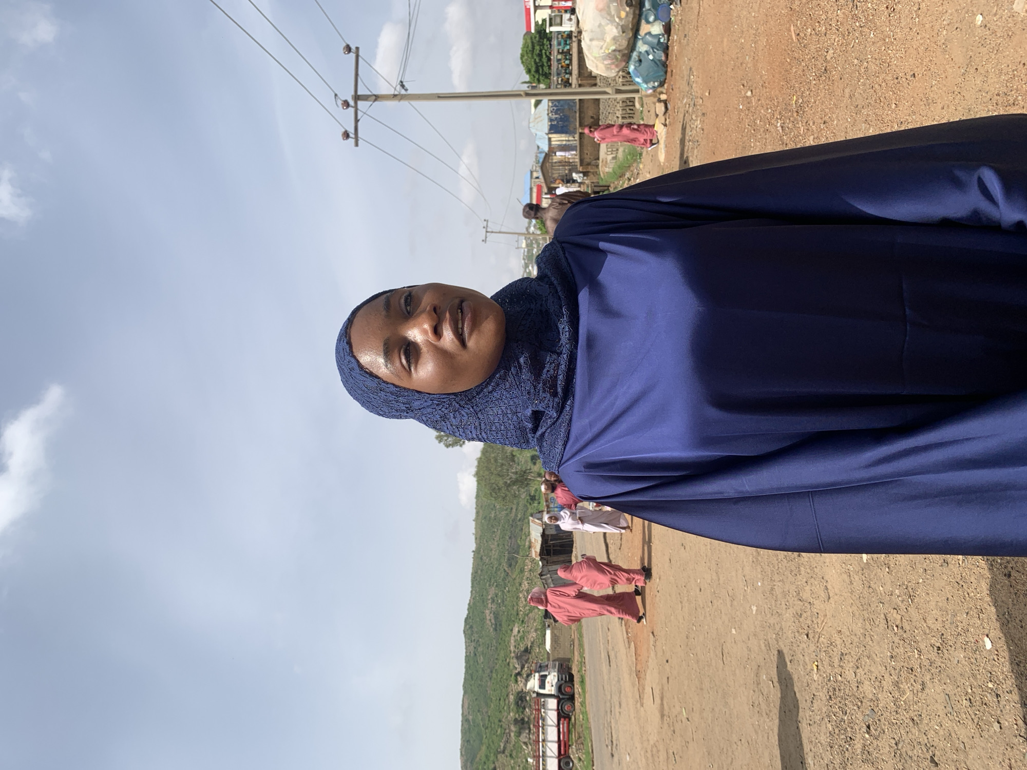 A woman wearing a blue hijab and dress stands outside in Naraguta Hausawa, Nigeria. She is the primary subject. In the background, other people wearing long garments are walking along a dirt road. Buildings are visible in the distance, along with a truck. Power lines are overhead. The sky is cloudy.