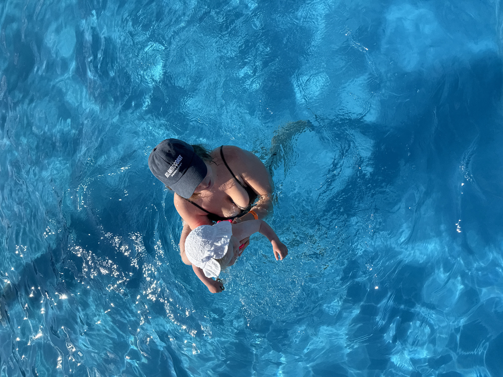 An aerial shot shows a woman in a black bikini holding a baby wearing a white sun hat in a bright blue swimming pool. The woman is wearing a dark gray baseball cap with "ATLANTIC COAST" printed on it. The baby is being held facing the viewer and is looking downwards. The water is clear with sunlight reflecting off its surface, creating shimmering patterns. The scene is set in a pool in the city of Koloksha, Russia.