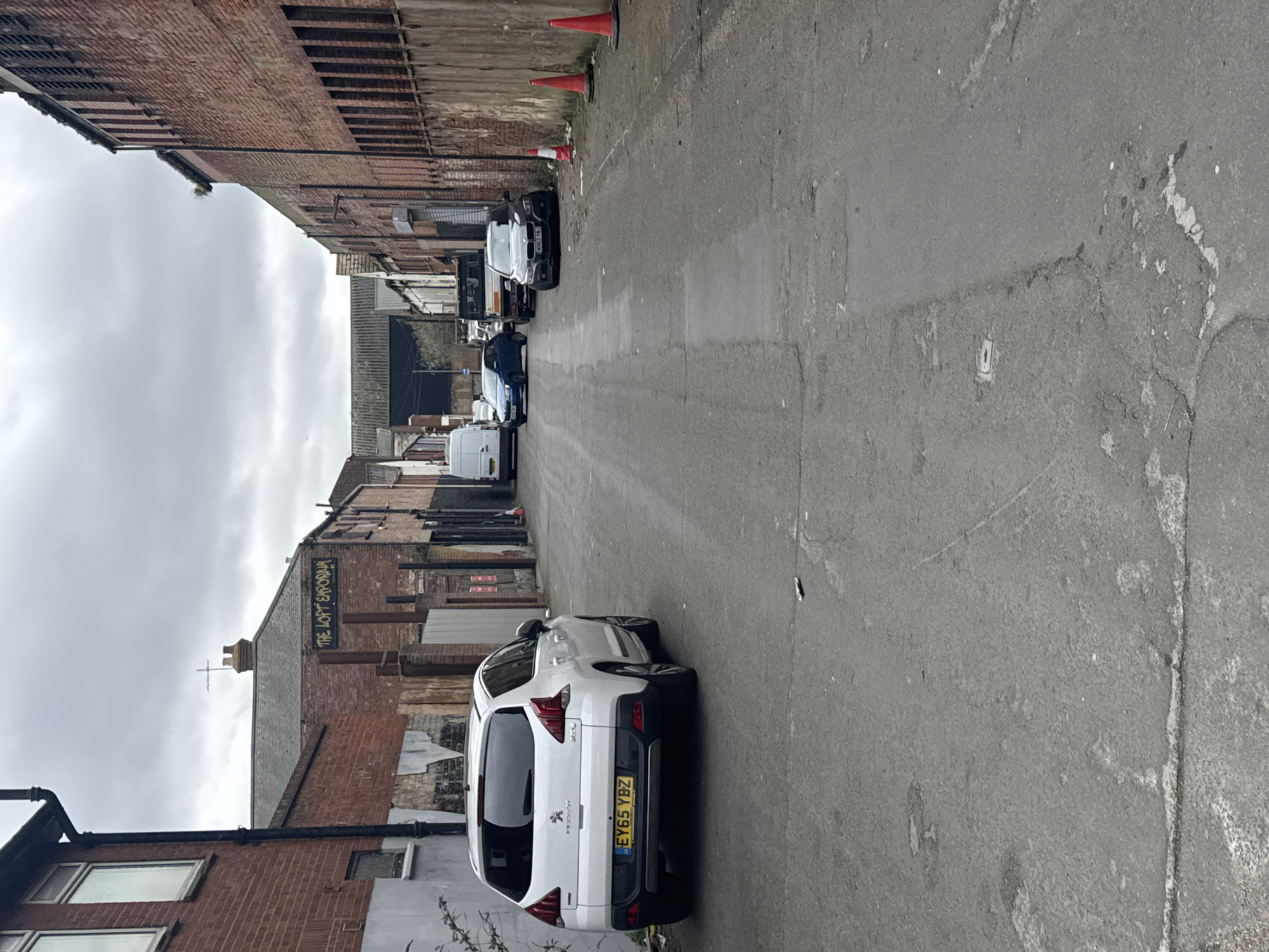 The image depicts a narrow alleyway in Gorleston-on-Sea, United Kingdom. The alley is paved with grey asphalt and flanked by buildings on either side.
On the left side, a red brick building with dark window frames and a sign that reads "THE LOFT EVELYN" is visible. Further down the alley on the left are several other buildings with varied brickwork.
Parked on the right side of the alley closest to the camera is a white SUV with the license plate "EY65 YBZ." Further down the alley, several cars are parked along both sides.
Traffic cones are placed sporadically along the left side of the alley further down. The sky is overcast, casting a flat, grey light over the scene. The alley is mostly empty, with no people immediately visible.
