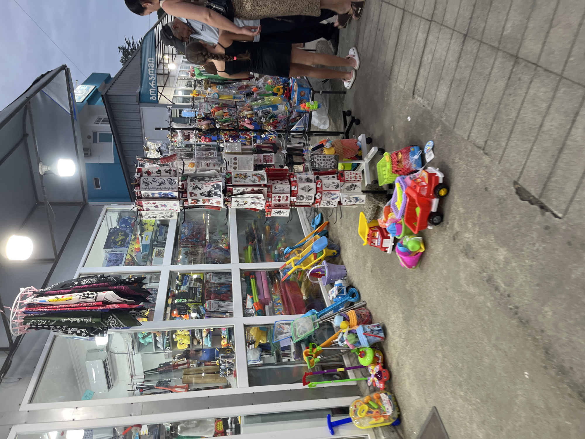 The image shows a street scene in Ureki, Georgia, featuring a retail stall filled with various items. The stall displays an array of products, including colorful children's toys such as toy trucks, shovels, buckets, and a toy suitcase with wheels. In the background, the stall also appears to sell other merchandise like bandanas, tattoos, and possibly fishing nets, with an air conditioning unit visible inside the store. Several people are present, including two women and an older man, some of whom are browsing the merchandise. The ground is paved with concrete, and the sky above suggests it is either dusk or an overcast day. The overall atmosphere is that of a bustling market or souvenir stand.