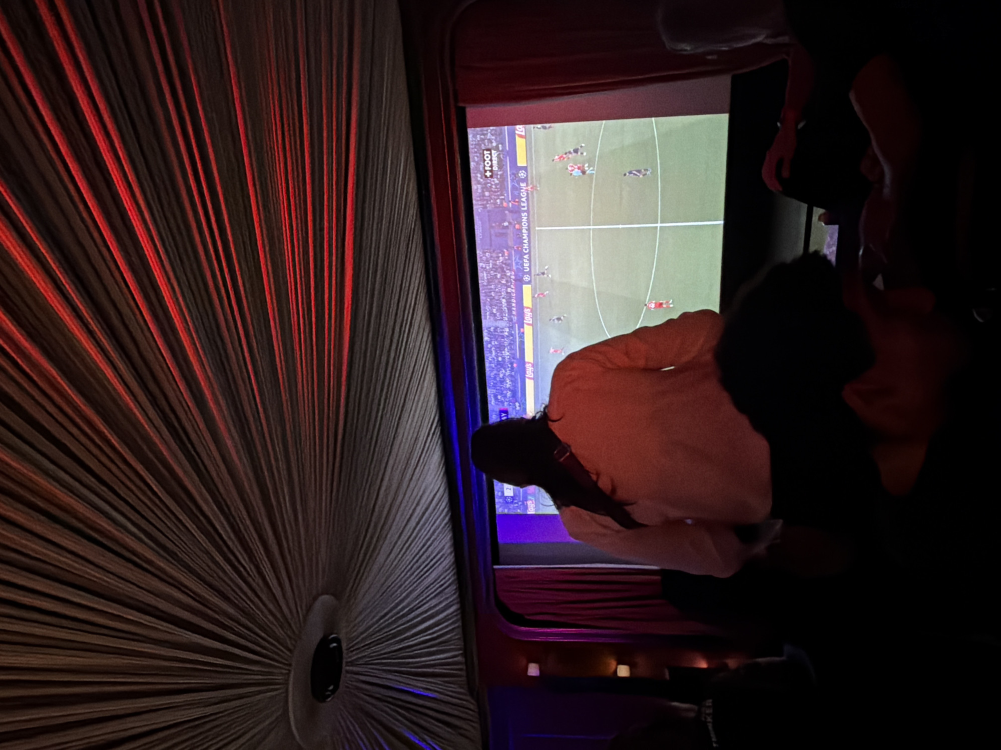 This media file captures a dimly lit indoor setting, likely a private viewing room, lounge, or bar, in Paris, France, during the evening or night. The primary focus is a large screen displaying a live soccer (football) match from the UEFA Champions League.
On the left side of the image, a textured wall or curtain features a striking radial pattern in dark grey or brown tones, illuminated by vertical streaks of red light emanating from a central circular point. This point contains a black, possibly metallic, fixture, which could be a speaker or a light source.
The large screen shows a green football pitch with players in red and dark kits. Visible text on the screen confirms it is a "UEFA CHAMPIONS LEAGUE" broadcast, featuring "+FOOT DIRECT" and a partially visible "Lays" sponsor logo.
Several people are present, mostly seen from behind or as dark silhouettes, engrossed in watching the match. One individual in the mid-ground, facing the screen, wears a light-colored top with a dark strap across their shoulder. The overall atmosphere is dark, with the bright glow of the screen providing the main illumination, complemented by the distinctive red ambient lighting on the left and some faint warm lights in the distant background.