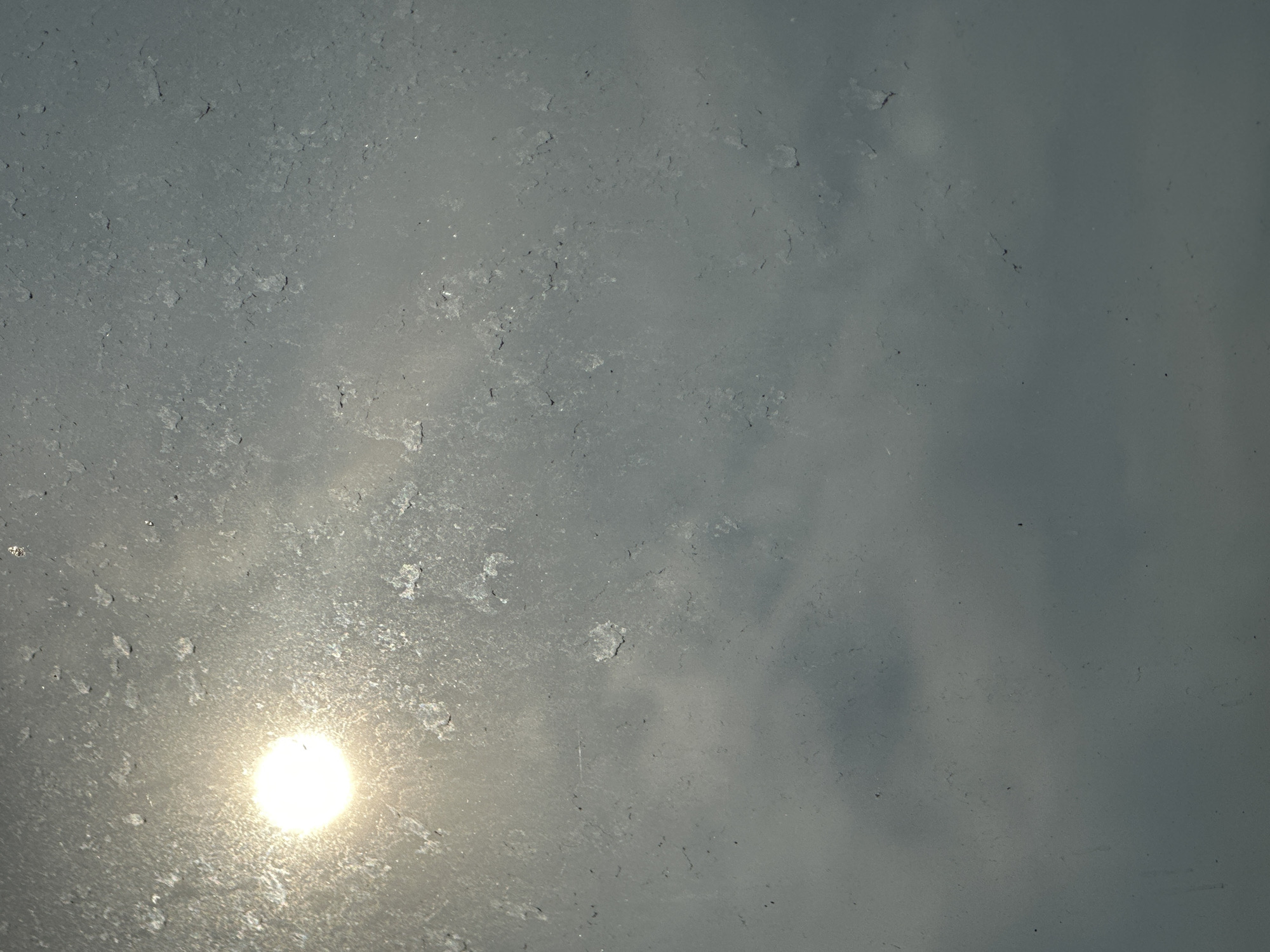 The image shows a bright, circular sun partially obscured by atmospheric haze and what appears to be frost or condensation on a windowpane. The sun is located in the upper left quadrant of the frame. The sky is a uniform gray, with some faint cloud formations visible. The foreground is dominated by the texture of the frosted or dirty window. There are no people, specific objects, or any discernible details of a city environment, including Podolsk, Russia. The image appears to be a close-up of a surface with the sun shining through.