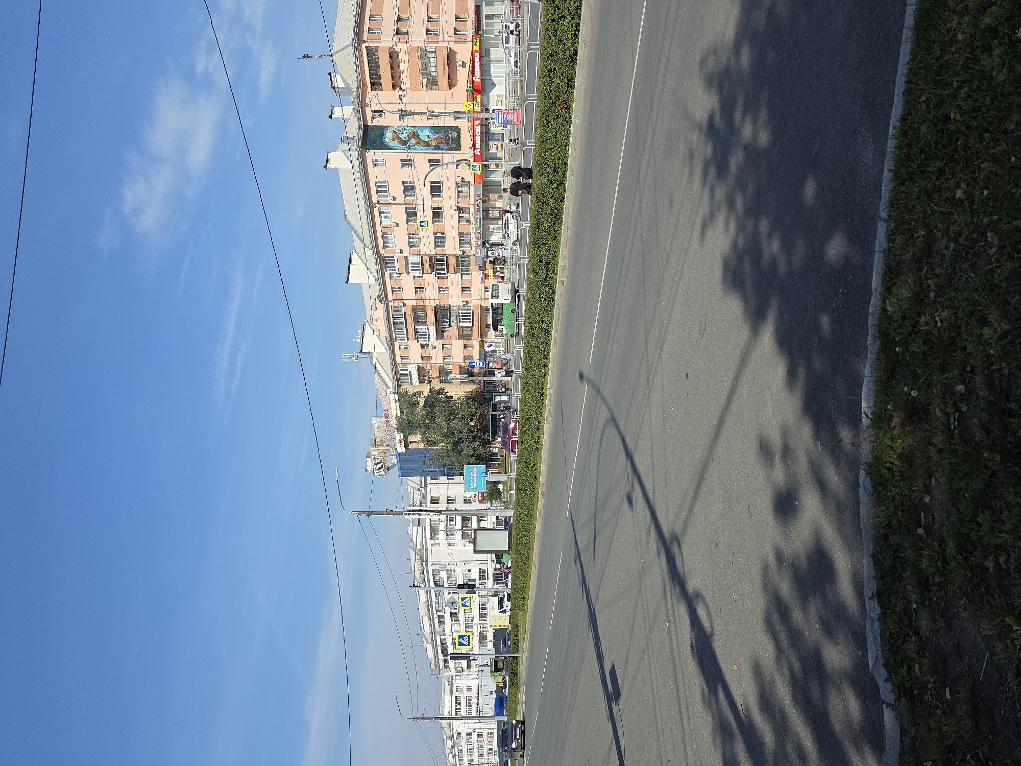 The image depicts a street scene in Chelyabinsk, Russia. On the right side of the frame, a multi-story residential building with peach-colored facades is prominent. The building features multiple windows, balconies, and a tiled roof. Several businesses are located on the ground floor, including shops with red and white signage that appears to read "Аптека" (Pharmacy). A large, colorful mural depicting a tree with a central figure is visible on the facade of this building. To the left of this building, a modern glass-fronted structure is visible, possibly an office building or commercial center, with a prominent sign that says "Оскар" and another smaller sign with Cyrillic text.
Across the street, a row of lighter-colored residential buildings stretches into the distance, characterized by numerous windows and balconies. Power lines crisscross the sky above the buildings. The street itself is a multi-lane asphalt road with clear lane markings. Several cars are visible on the road, including a black sedan in the left lane and other vehicles further down. A green bus is also seen in the distance. Pedestrians are present on the sidewalk in front of the businesses, with two individuals in dark clothing standing near the sidewalk.
The environment is characterized by a clear blue sky with some wispy white clouds. The street is lined with a manicured green hedge. Shadows of trees and power poles are cast across the asphalt. The foreground shows a patch of grass and soil along the edge of the road. The overall impression is that of a sunny day in an urban setting.