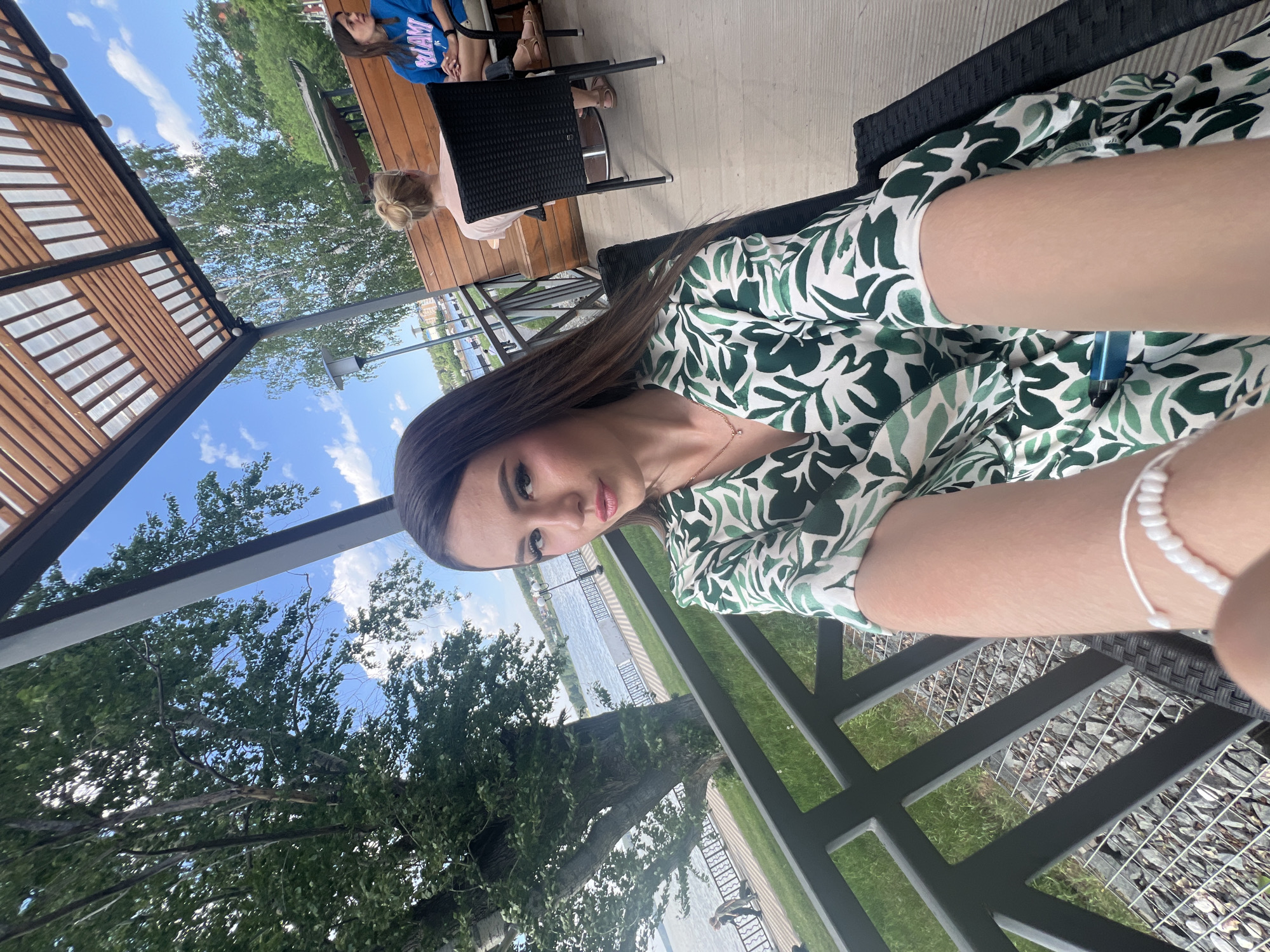 A woman is sitting at an outdoor table in what appears to be a restaurant or cafe, possibly in the city of Votkinsk, Russia. Behind her, a body of water is visible, likely a river or lake. A tree is partially visible on the left, and there are other people seated at tables in the background. She is wearing a patterned green and white dress and a white beaded bracelet is visible on her arm.