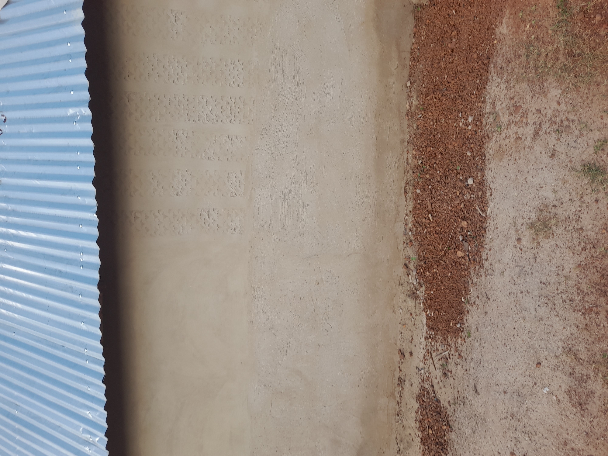 The image shows a wall of a building with a corrugated metal roof in Ibi, Nigeria. The wall is made of light-colored cement and has a textured pattern of small, repeating wave-like shapes in vertical columns. The roof is blue and appears to be made of corrugated metal sheets fastened with nails or screws. The ground in front of the wall is covered with dirt and small reddish-brown stones, with some sparse dry grass. There are no people or animals visible in the image. The scene appears to be an exterior view of a building under construction or recently built.