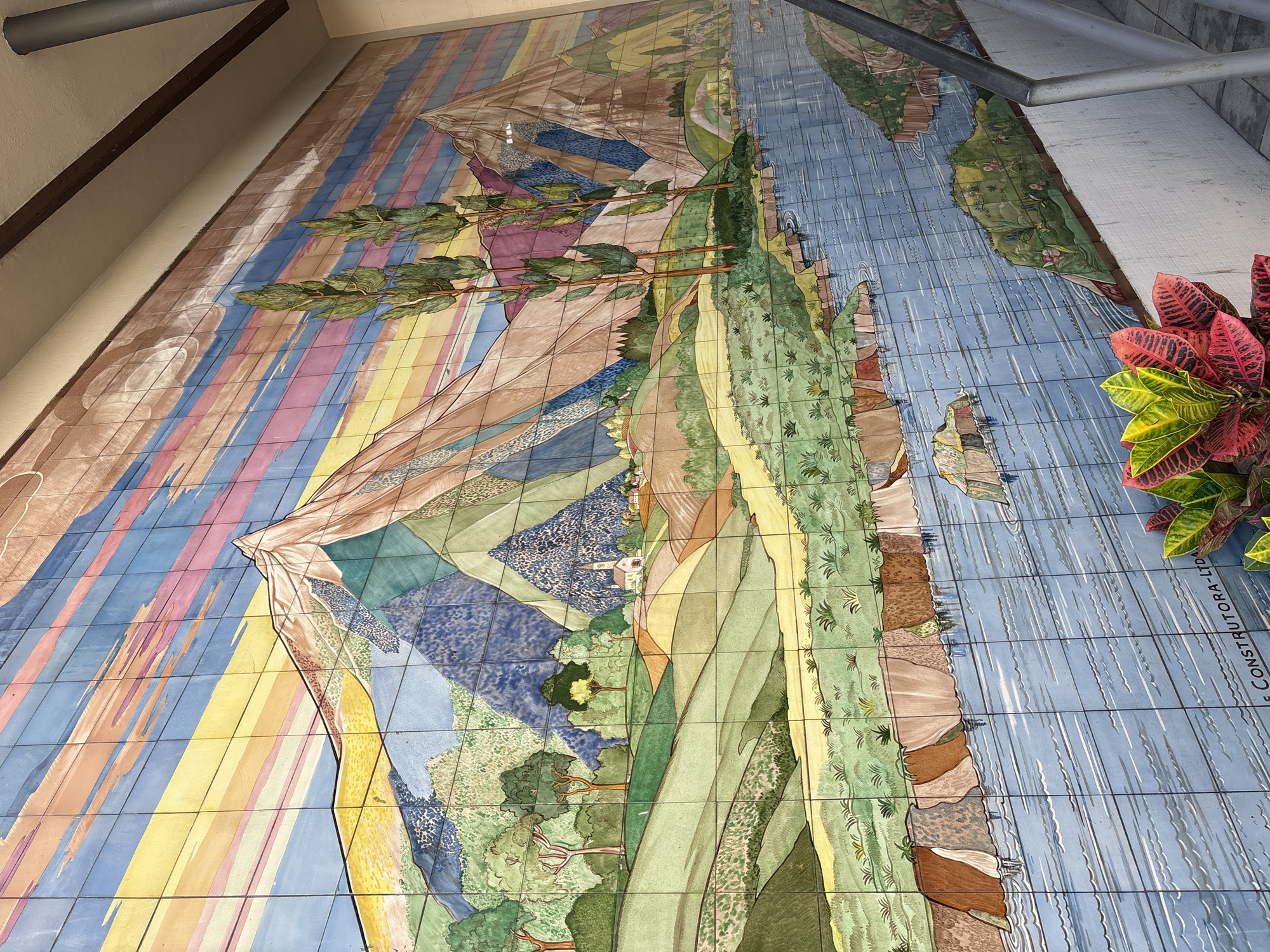 This is a tiled mural depicting a stylized landscape of mountains, rolling hills, trees, a river, and a village with a church. The sky is rendered with horizontal bands of color, suggesting a sunset or sunrise. The mural is installed on an exterior wall of a building, possibly under an awning or overhang, as indicated by the shadow cast from the upper right. A metal railing runs along the lower right edge of the image, bordering a set of stairs. In the foreground, a potted plant with vibrant red and green leaves is visible. The mural is signed at the bottom with "E CONSTRUTORA - LTDA". This mural is located at the Colégio São Luís in São Paulo, Brazil, on the facade of the "Anexo" building.