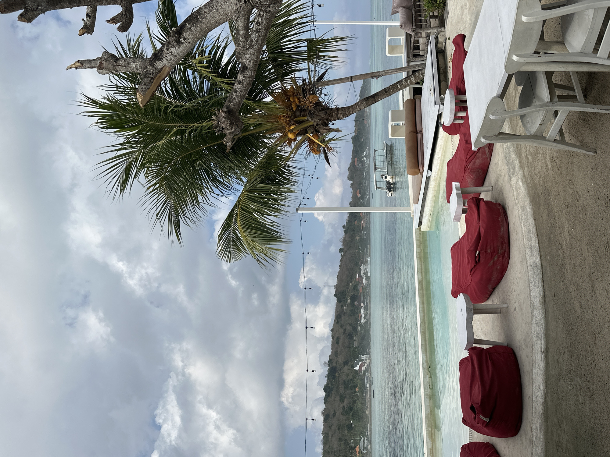 The image depicts a relaxed outdoor seating area near a swimming pool, with a view of the ocean and a distant coastline. The seating consists of several large, red beanbag chairs and white tables. In the foreground, a white wooden table with chairs suggests a dining area. Beyond the pool, a wooden platform with cushioned seating is visible. A boat with the name "SUNDAY BEACH" is anchored in the calm, turquoise water. Lush green palm trees, one laden with coconuts, dominate the right side of the frame, with their fronds casting shadows. The sky is partly cloudy with a mix of white and grey clouds. The coastline in the distance is lined with trees and some buildings. This scene is consistent with a beachside resort or restaurant in Nusa Penida, Indonesia, specifically likely at a venue like Sunday Beach Club.