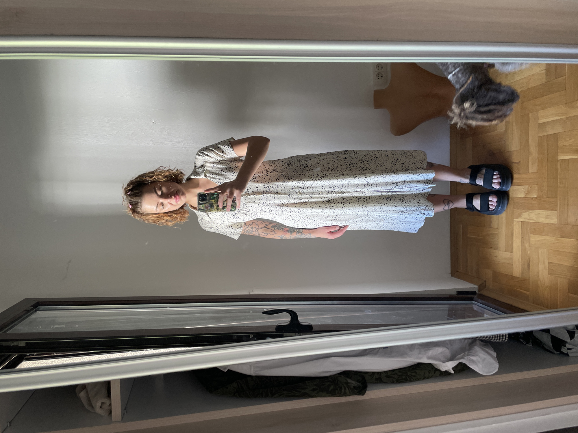 A woman is taking a mirror selfie. She is wearing a white dress with small black spots and black sandals. She has curly blonde hair and tattoos on her arm. The background includes a white wall, wooden floor, and a mannequin head. The mirror reflects a closet with clothes inside. The location is likely indoors and cannot be precisely pinpointed to Boreti, Montenegro based on the provided image.