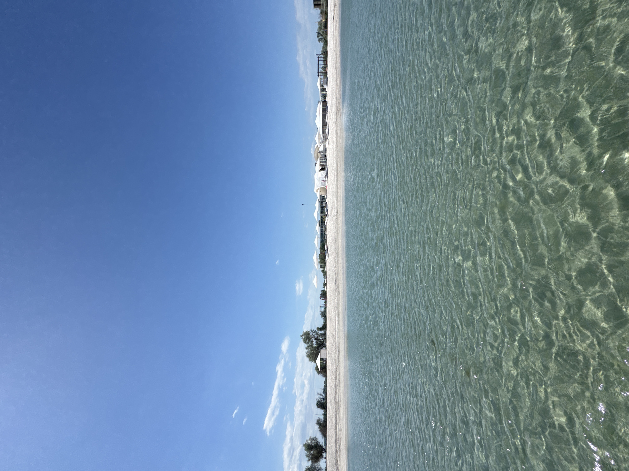 The image depicts a beach scene, likely near a resort. Clear turquoise water fills the right half of the frame. A white sand beach runs horizontally across the middle, separating the water from a clear blue sky with a few scattered clouds on the left. Along the beach, there are white beach umbrellas and some sparse vegetation. The location is not discernible from the image.