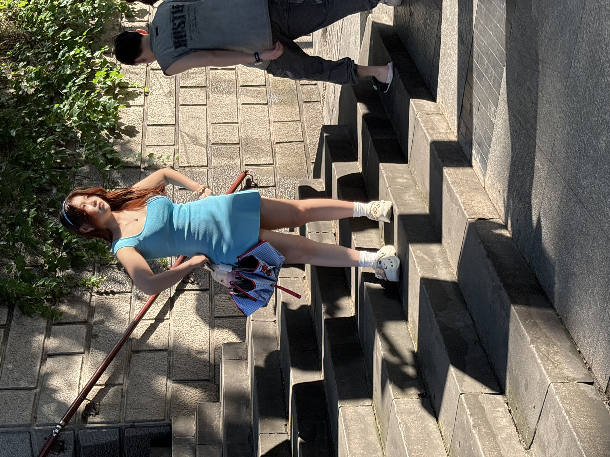 In Yuzhong District, China, a young woman with long, auburn hair, wearing a light blue sleeveless dress and white Croc-style shoes, stands on a stone staircase. She has her hands on her hips and is holding a small, folded blue and white checkered umbrella with red trim. Next to her, to her right, a person wearing a grey sleeveless shirt with "HUSTLE" printed on it and dark pants is walking up the stairs. The stone wall behind them is covered in green ivy, and sunlight casts shadows on the stairs and surrounding pavement.