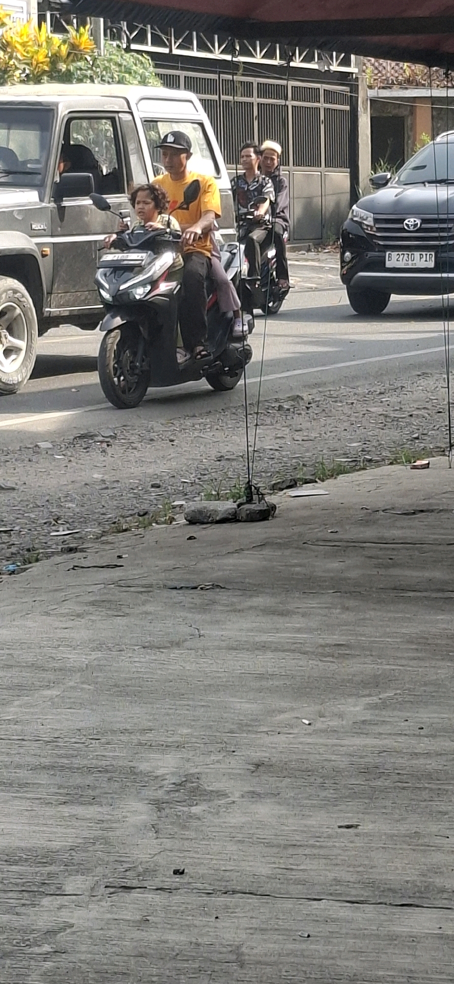 In Rancagoong, Indonesia, a man in a yellow t-shirt and a black baseball cap rides a black scooter. He is accompanied by a young child with curly hair who is seated in front of him. Behind the man, two other individuals are also riding on scooters. The first, a man wearing a patterned shirt and a white peci (skullcap), is on a scooter with a second passenger wearing a dark outfit and a peci. The scooters are on a paved road, with a gray SUV in the background. The environment appears to be a street scene, possibly in a suburban area, with some greenery and a building visible behind the vehicles.