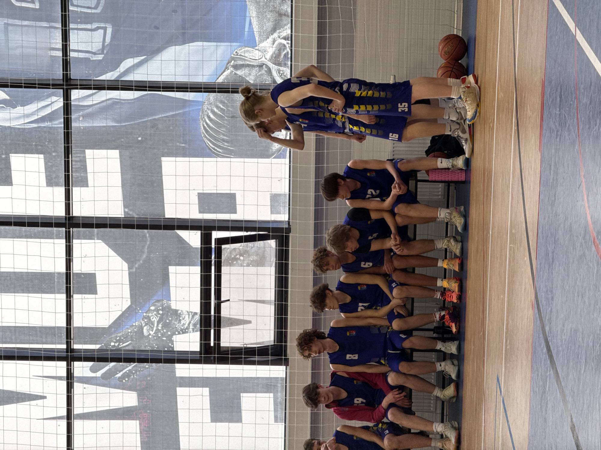 A sports team sits on a bench indoors in Votkinsk, Russia. The players wear blue and yellow uniforms and basketball shoes. Two basketballs rest on the floor. A window is behind the team.