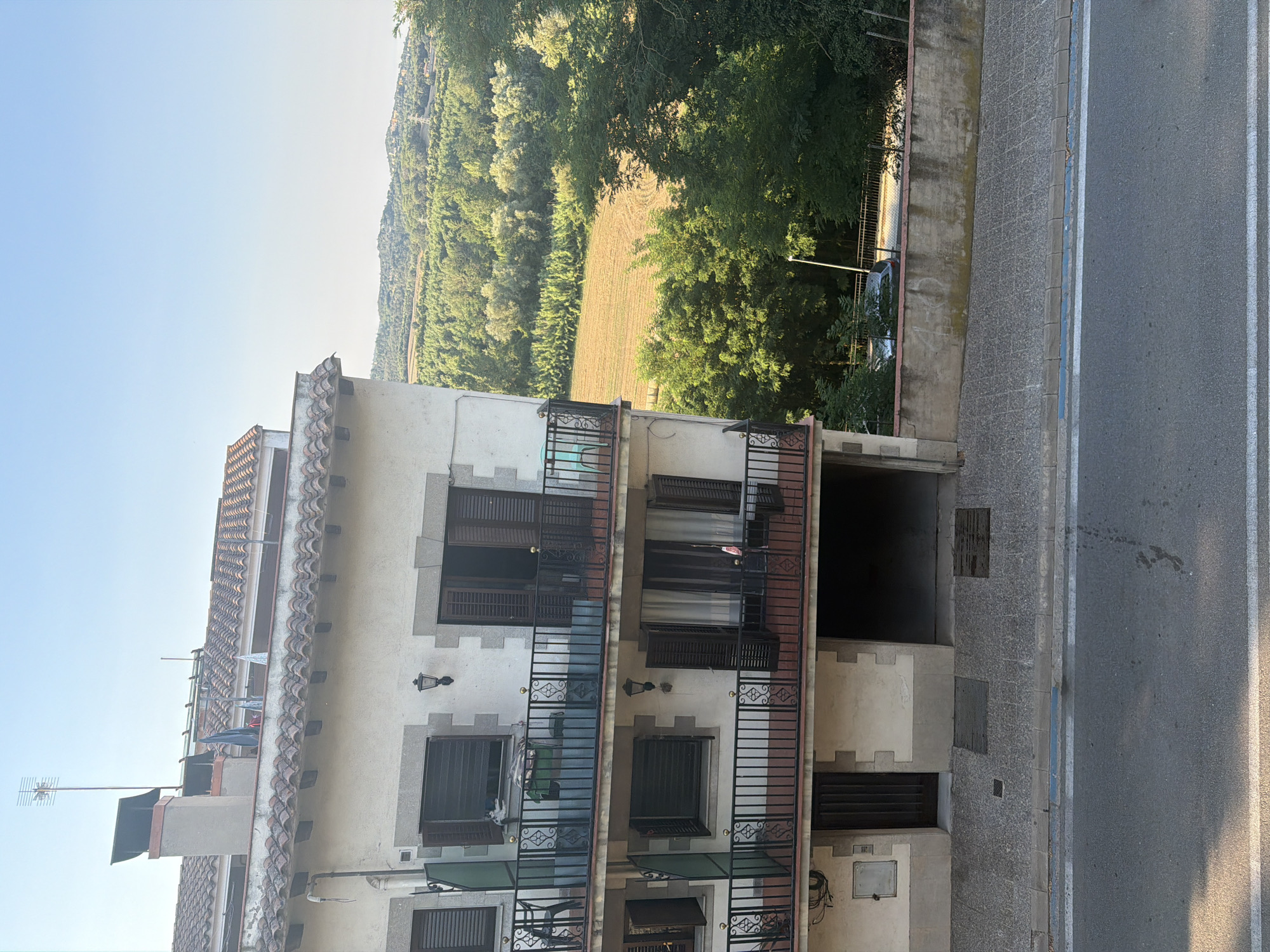 The image captures a view from an elevated position looking down onto a three-story building in Hostalric, Spain. The building's exterior is primarily white with stone accents around the windows and balconies. It features multiple balconies with black metal railings, some with chairs and laundry visible. The top of the building is adorned with a red tiled roof and a chimney.
Beyond the building, a lush green landscape unfolds. A field with agricultural patterns is visible, surrounded by dense trees and rolling hills that recede into the distance under a clear blue sky. A road runs in front of the building, with a sidewalk and a low concrete wall separating it from the property. A light-colored car is parked beyond the wall, partially obscured by trees. The scene suggests a residential area integrated with the natural environment.