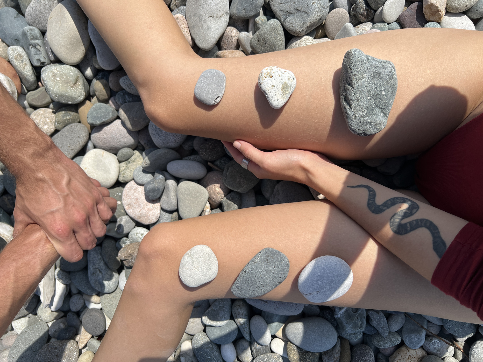The image shows a top-down view of two people sitting on a pebble beach, likely in Batumi, Georgia, given the presence of a snake tattoo on one individual's arm, a common motif in Georgian folk art and symbolism. The hands of two individuals are clasped together at the top of the frame. In the foreground, the legs of two people are positioned. On the left leg, four stones are placed: a light grey oval stone above the knee, a darker grey, flat, elongated stone below the knee, and two lighter grey stones, one oval and one more irregular, further down the leg. The right leg has three stones: a small, rounded grey stone above the knee, a heart-shaped white and tan stone below it, and a dark grey, irregular stone lower down. A snake tattoo is visible on the left forearm of one individual, which is resting on their leg. The environment consists of a dense scattering of various sized and colored pebbles. The lighting suggests a sunny day with distinct shadows cast by the stones and legs.