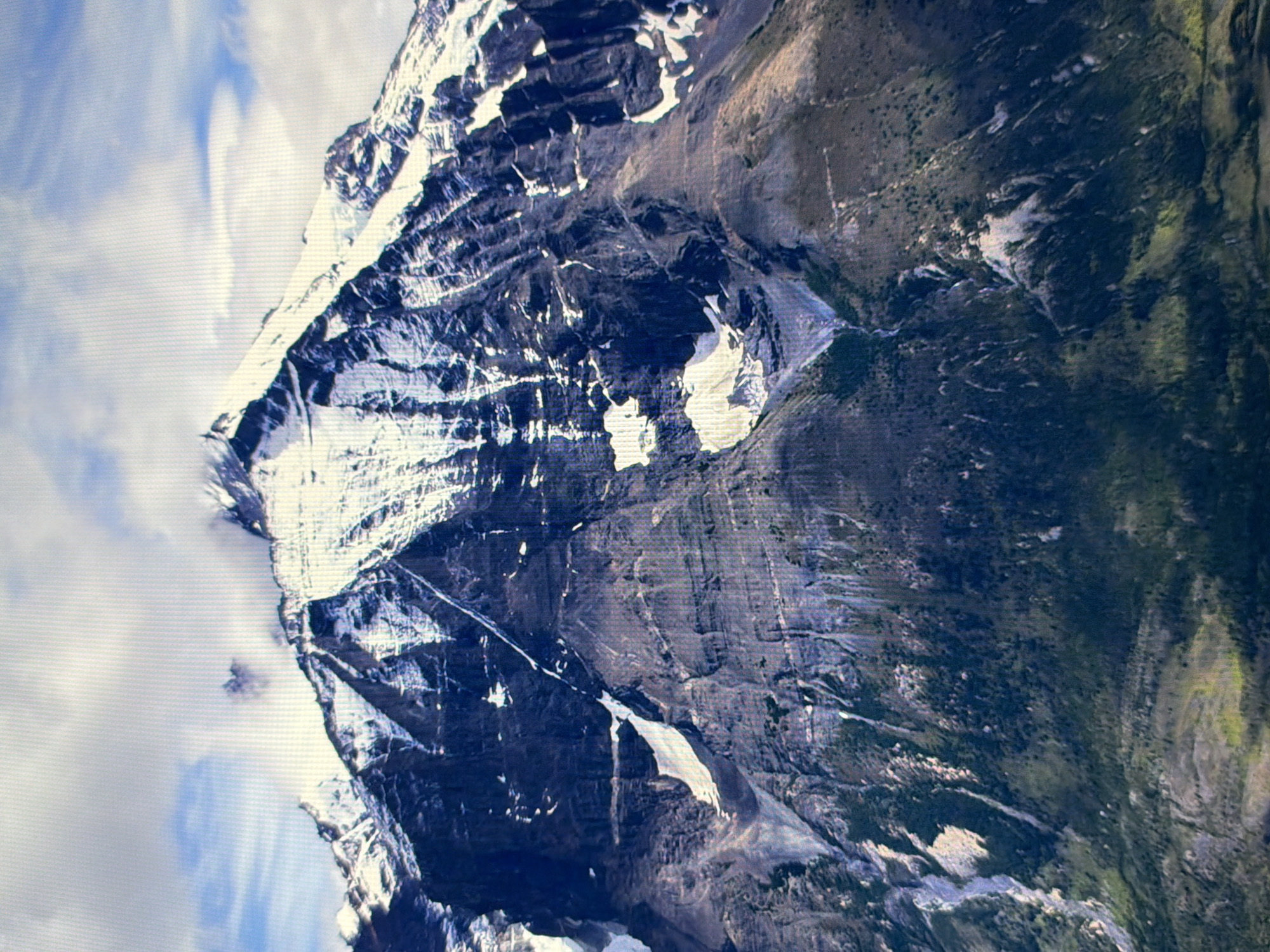 The image is a wide shot of a majestic, snow-capped mountain range under a cloudy sky. The mountain is steep and rugged, with dark rock faces streaked with white snow and ice. Patches of green vegetation are visible on the lower slopes, suggesting a transition from alpine to subalpine environments. The clouds are a mix of white and gray, with some areas of blue sky peeking through. The lighting suggests it is daytime. There are no people, structures, or any indication of a city in the image. The scene depicts a natural, mountainous landscape. There is no visible connection to the city of Kyiv, Ukraine.
