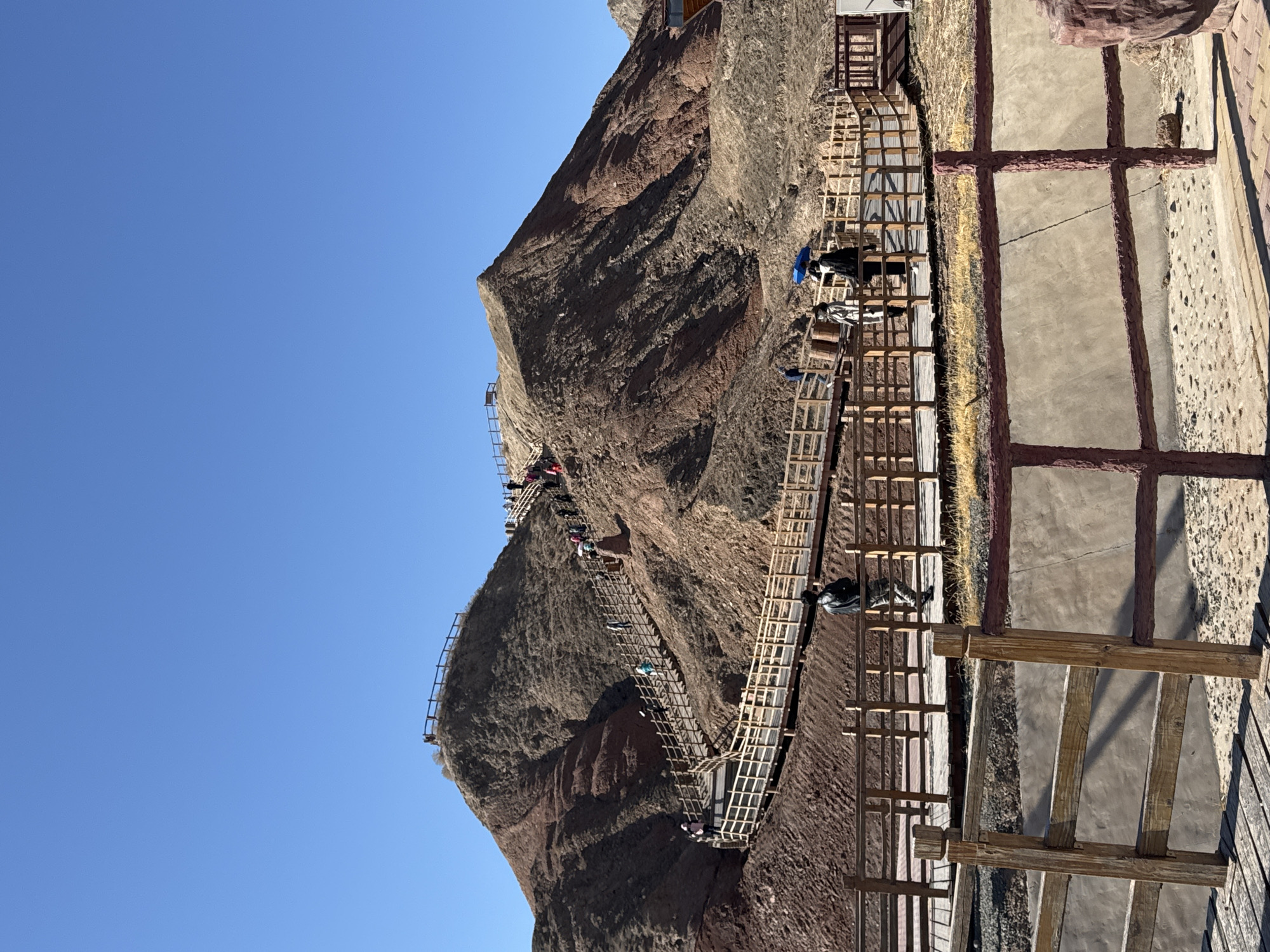The image displays a steep, reddish-brown mountainous landscape under a vast, clear blue sky. An extensive system of wooden walkways and stairs is meticulously built into and alongside the mountain face, designed to facilitate tourist access. Numerous people, appearing to be hikers or sightseers, are visible along these pathways, actively ascending the mountain.
The scene depicts a rugged, arid environment characterized by prominent rock formations with distinct red and brown hues. The wooden pathways feature railings for safety and appear to be well-maintained, indicating a developed tourist attraction. In the mid-ground, one individual is seen holding a bright blue umbrella, possibly for sun shade, while others carry small bags or backpacks.
The bright, direct sunlight and the deep blue sky suggest a clear, sunny day, likely during the morning or early afternoon. The overall setting points to an outdoor recreational area or a scenic geological park, consistent with the location context of Nantai, China. There is no visible text in the image.