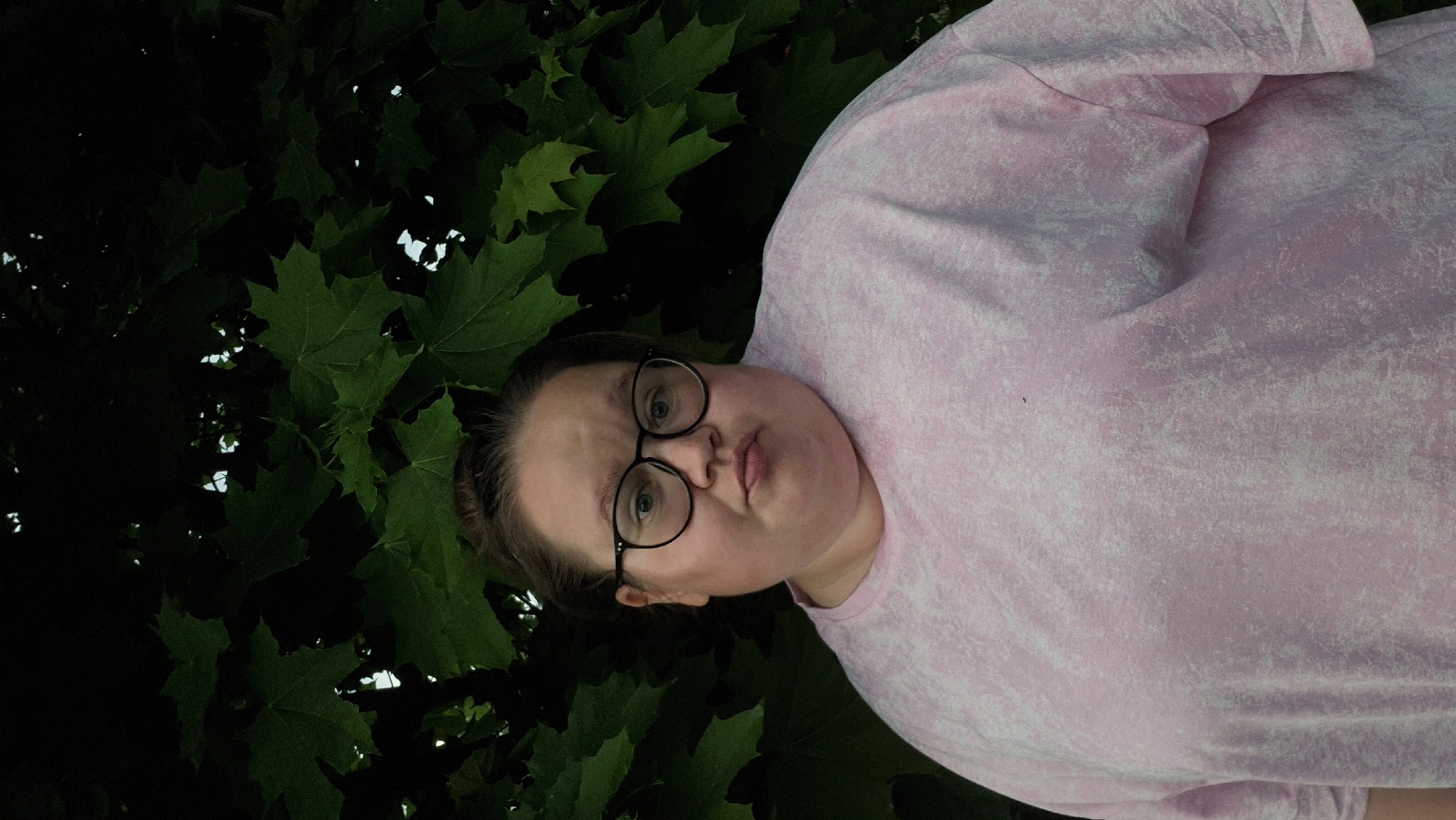 A portrait of a person with round glasses, wearing a pink t-shirt, set against a backdrop of green leaves. The image appears to be rotated. The location, Chaikovsky, Russia, cannot be determined from the provided image.