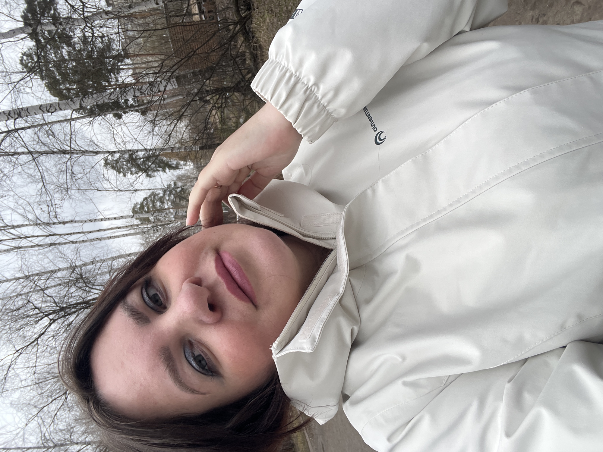 A woman in a white jacket is taking a selfie outdoors, with trees in the background, likely in Perm, Russia.
