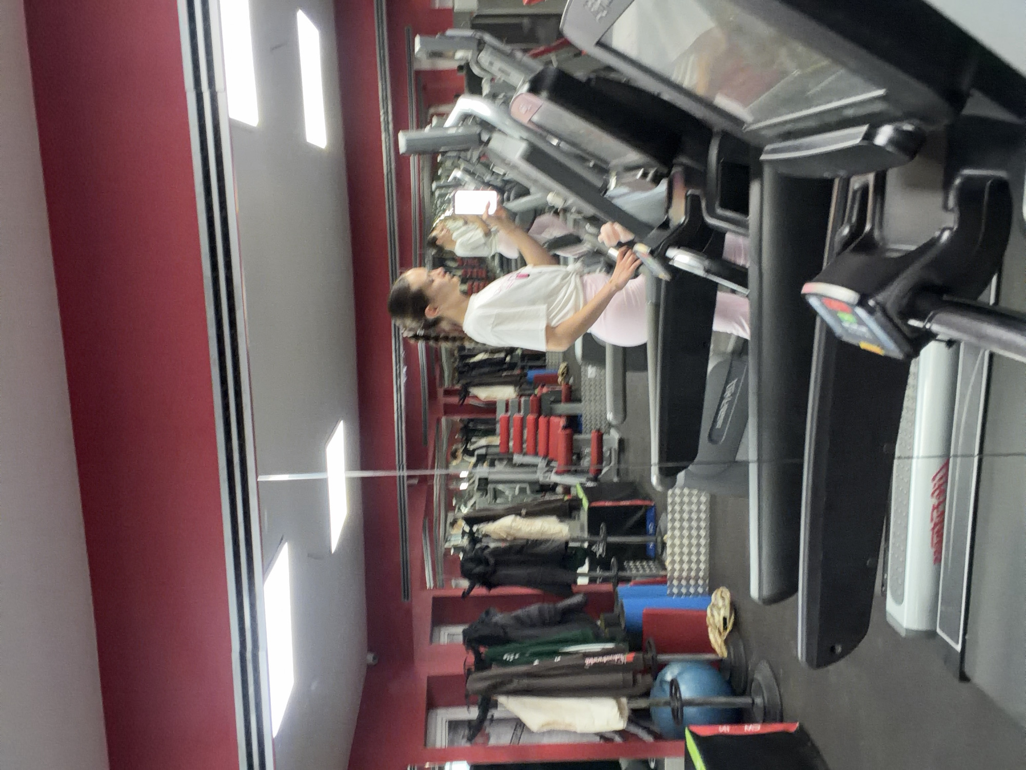 A young woman in a white t-shirt and light pink leggings is on a treadmill in a gym. She is holding a smartphone and appears to be taking a picture or video. Behind her are rows of other treadmills and various workout equipment. A wall with a mirror reflects the scene and the gym's interior, revealing a red wall and linear lighting fixtures on the ceiling. The location is likely in Sofia, Bulgaria, although this cannot be confirmed from the image alone.