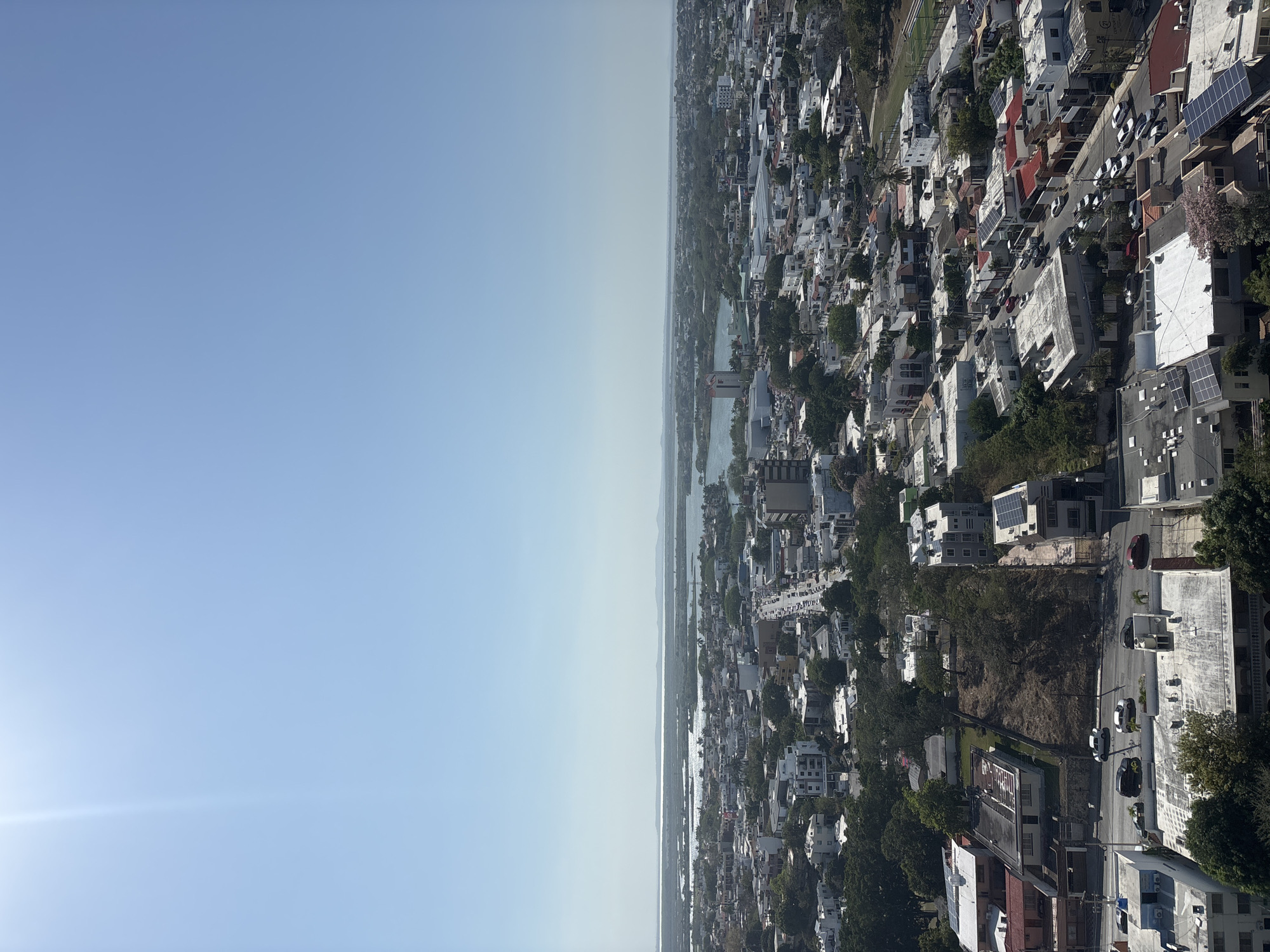 The image shows an aerial view of Tampico, Mexico. The left side of the image shows a clear, light blue sky. The right side shows a densely populated area with a mix of residential and commercial buildings. The buildings are mostly white or light-colored, with some having red roofs. Solar panels can be seen on some of the buildings. Narrow streets wind through the urban landscape, with cars visible on some of them. Trees are interspersed among the buildings, adding greenery to the scene. In the distance, there appears to be a body of water, possibly a river or a lake. The image seems to be taken from a high vantage point, likely from a tall building or a drone, providing a panoramic view of the city.