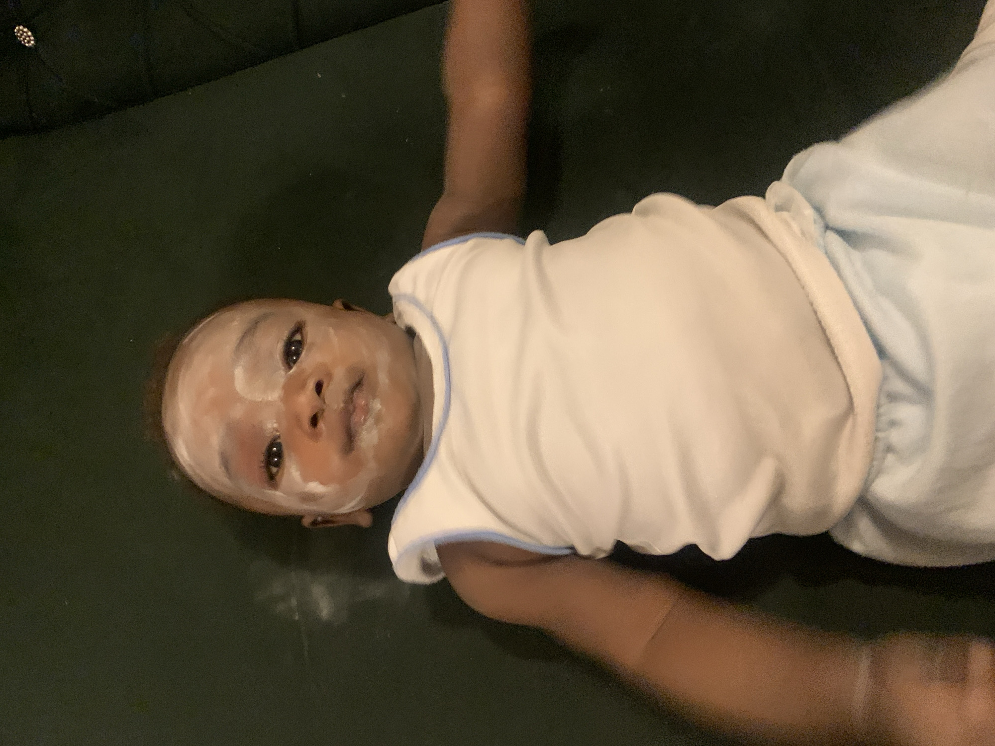 A baby lies on a dark green surface. The baby is wearing a white shirt with blue trim and light blue bottoms. There is a white substance smeared on the baby's face. The location is assumed to be in Barakin Akawo, Nigeria.
