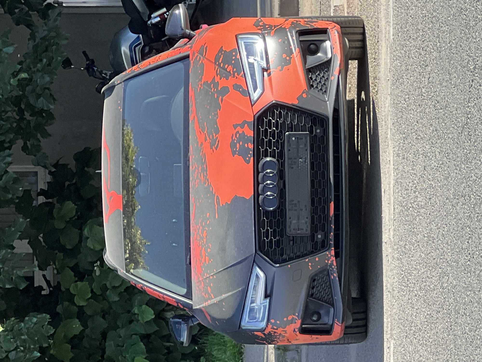 The image displays an Audi car, viewed from a frontal-right, slightly elevated perspective, parked on a street during bright daylight. The car features a striking custom paint job or wrap, characterized by a matte dark grey or black base extensively covered with vibrant orange, irregular splatter patterns across the hood, front fenders, and bumper.
The front of the car is highly detailed, showing a prominent, large, hexagonal grille with a honeycomb mesh pattern and the iconic four-ring Audi logo in the center. Modern LED headlights with distinctive internal light signatures are visible on both sides of the grille, alongside aggressive air intakes integrated into the front bumper. A dark, rectangular panel, likely a blank license plate holder, is present within the grille, but no readable text is visible on it.
Behind the car's roof, on the upper left side, parts of a motorcycle or scooter are visible, including handlebars and a mirror. Lush green foliage, possibly from a bush or tree, fills the background on the left and reflects in the car's windshield. The car is situated on an asphalt road surface, adjacent to a light grey curb or sidewalk on the right. Strong sunlight illuminates the scene, creating clear shadows and highlighting the car's unique finish. The setting appears to be an outdoor urban or suburban environment, consistent with a street parking scenario in Krasnodar, Russia.