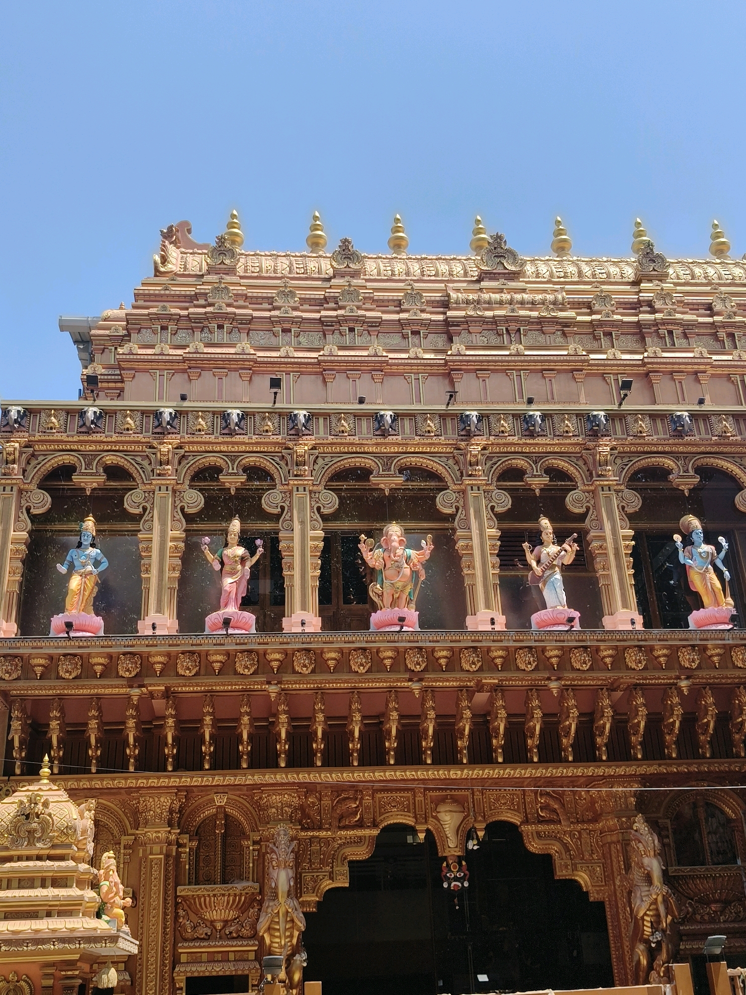 The image shows a close-up, low-angle exterior view of a vibrantly decorated Hindu temple, likely located in Thiruvananthapuram, India. The temple architecture is elaborate, featuring intricate gold detailing, carved figures, and a pastel pink and gold color scheme. Several colorful statues of deities are prominently displayed on an upper level, each standing on a pink lotus-shaped base. From left to right, these appear to be Vishnu, Lakshmi, Ganesha, Saraswati, and another representation of Lakshmi. Above these figures, a series of ornate arches and decorative elements, including what appear to be carved elephants and lions, are visible. The temple roof is adorned with golden finials and intricate carvings. The sky above is a clear, bright blue. The lower part of the image shows more detailed gold carvings and archways, with a dark, recessed entrance visible in the center. The overall impression is one of grandeur and religious devotion.