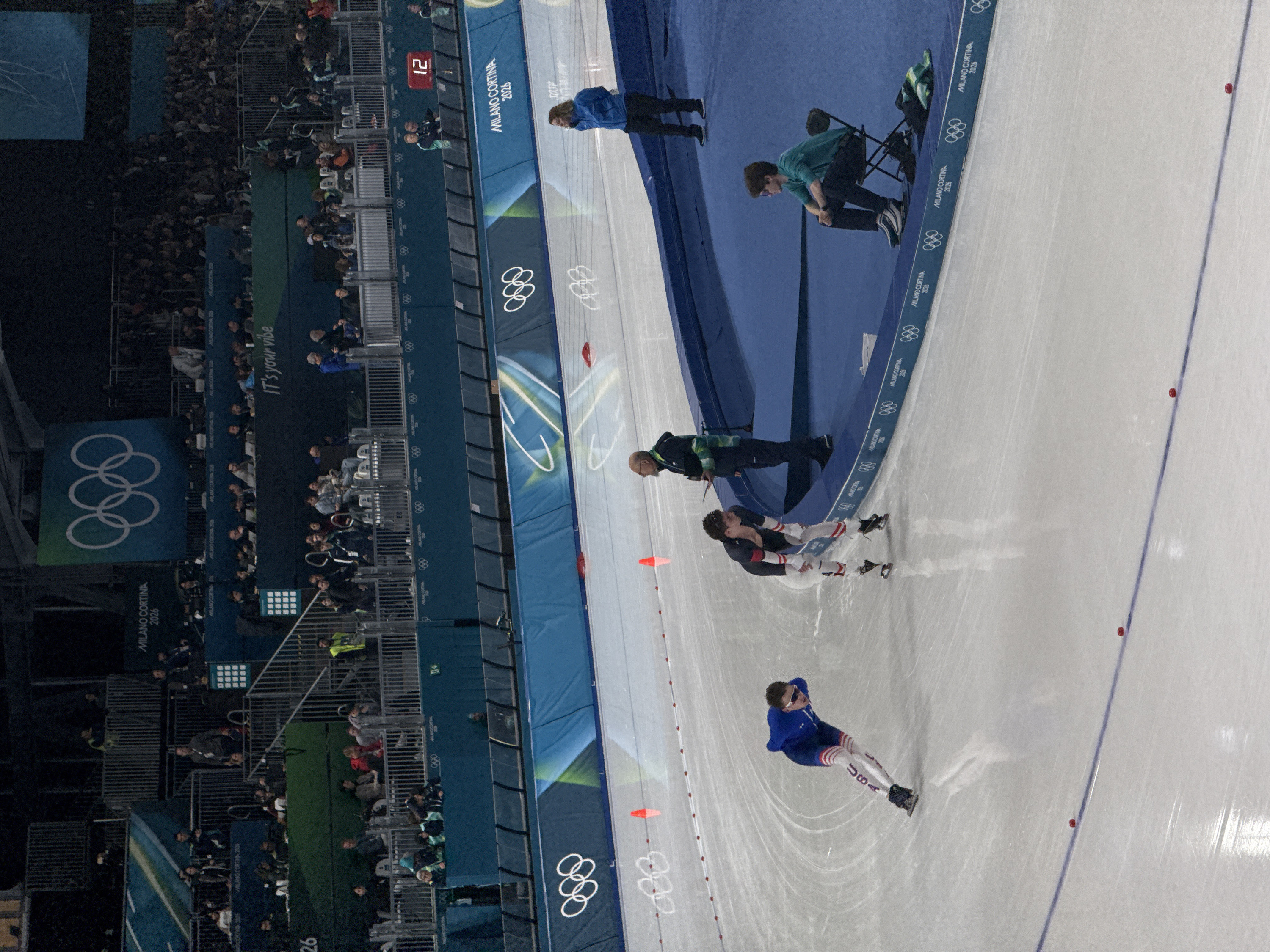 This image captures an indoor speed skating event or practice, likely at an arena preparing for or hosting the Milano Cortina 2026 Olympic and Paralympic Games. The perspective is from an elevated position in the stands, looking down onto the ice track.
In the foreground, a speed skater wearing a blue and white suit with "USA" prominently displayed on the left thigh is actively skating on the ice. This skater appears to be making a turn, with their body angled and arms tucked, demonstrating the dynamic posture of speed skating. Further down the track, a second speed skater in a white and black/red suit is also on the ice, possibly waiting or having just completed a segment.
Along the edge of the ice track, several individuals, likely coaches or officials, are present. One person in a blue jacket stands near the barrier, observing. Another person in a teal/green long-sleeved shirt is seated on a black folding chair by the barrier, seemingly focused on the track. A third individual, wearing a dark jacket with a green and striped bib or vest, stands near the second skater.
The ice surface is marked with a blue line and several small red cones or markers, indicating the track layout. A blue barrier separates the ice from the spectator area. This barrier is adorned with multiple white Olympic rings logos and the text "MILANO CORTINA 2026" repeated along its length.
To the left of the ice track are spectator stands, filled with numerous individuals, although many are blurred or in shadow due to the distance and lighting. Visible within the stands are metal railings and various banners. A large banner with the Olympic rings in white against a blue and green background is prominent. Another banner features the text "IT's your vibe". A red digital display board shows the number "12". The overall lighting suggests an indoor setting with artificial illumination, common for ice sports events.