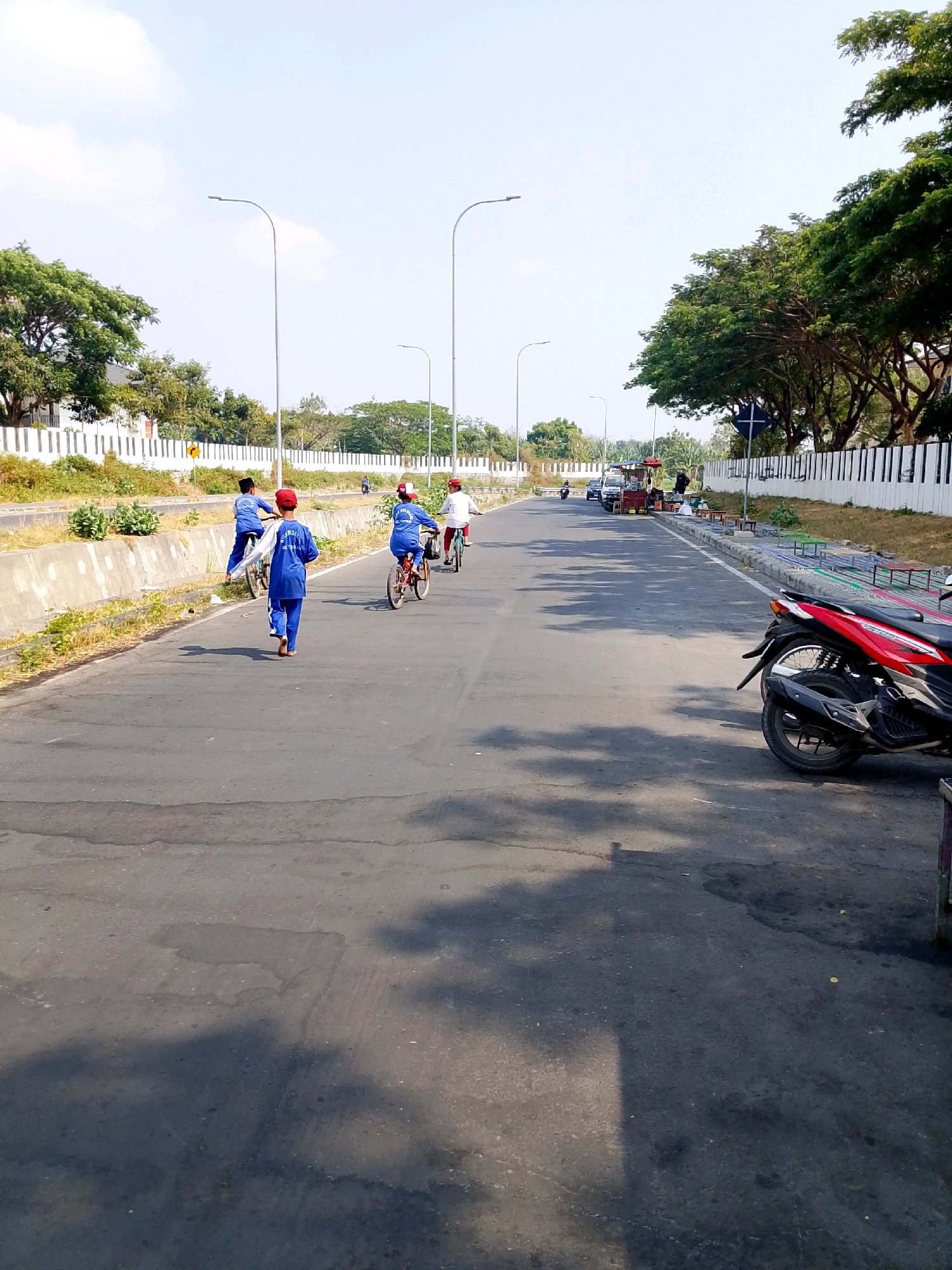 In Bulusari, Indonesia, a paved road stretches into the distance under a clear sky. On the left side of the road, a concrete barrier separates the asphalt from a grassy embankment lined with bushes. Further left, a white fence marks the boundary of a residential area with houses and mature trees. Several young individuals on bicycles are visible. Three boys in blue uniforms are on the left side of the road, two on bicycles and one walking. Another child in a red hat and blue uniform is riding a bicycle further ahead. On the right side of the road, a red and black motorcycle is parked. In the distance, a food cart and several people are gathered. Streetlights are spaced along the road. The scene depicts an everyday street activity with people commuting and enjoying leisure time.