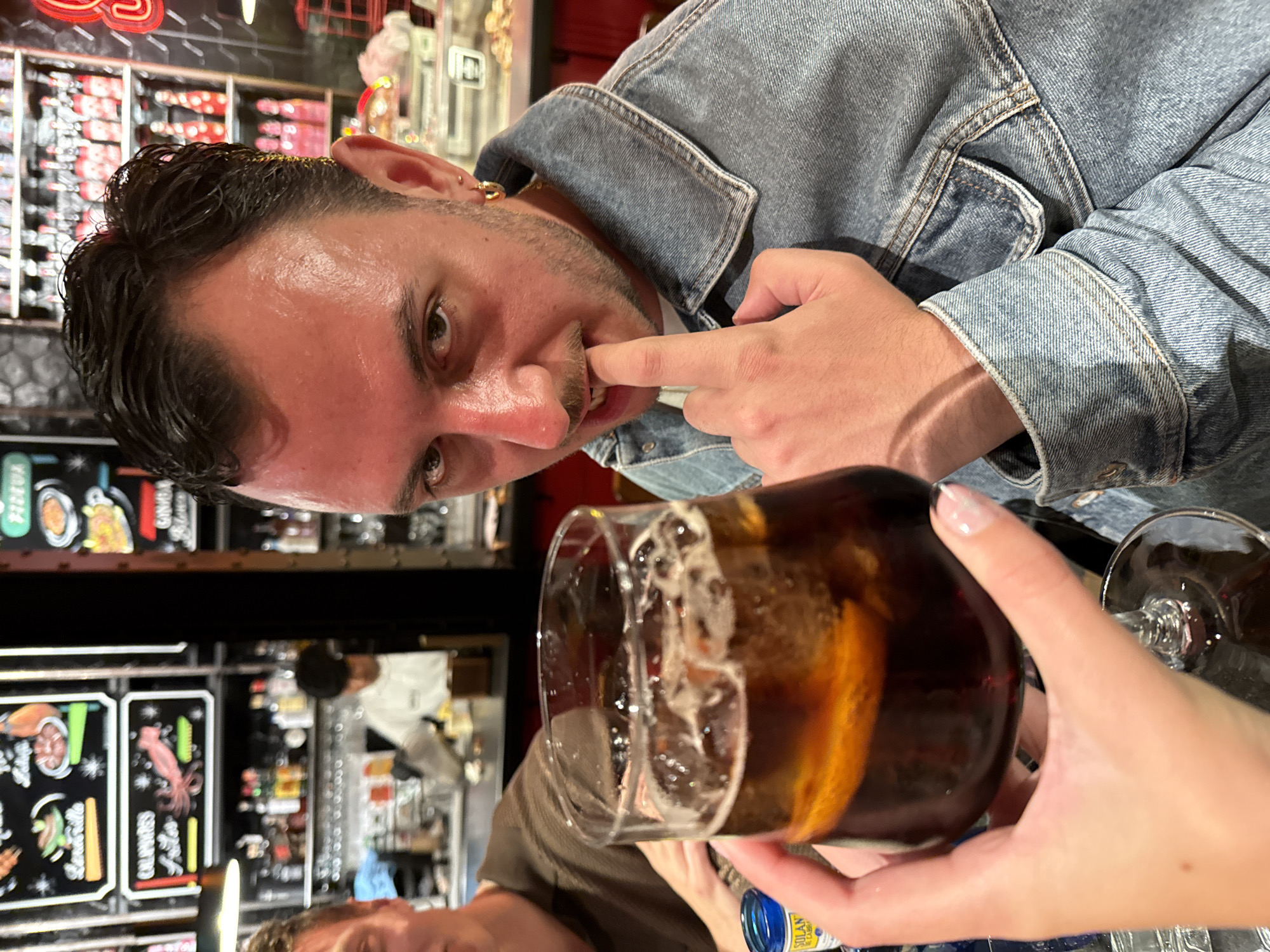 A medium shot depicts a man wearing a light blue denim jacket, with his right index finger partially inside his mouth. He is slightly angled towards the camera, with his eyes directed at the lens. The background appears to be a restaurant, specifically a bar area with dark furniture. Menu boards or signs are visible on the wall, with various food items listed. A hand holding a drink is near the bottom of the shot.
The location is Madrid, Spain.