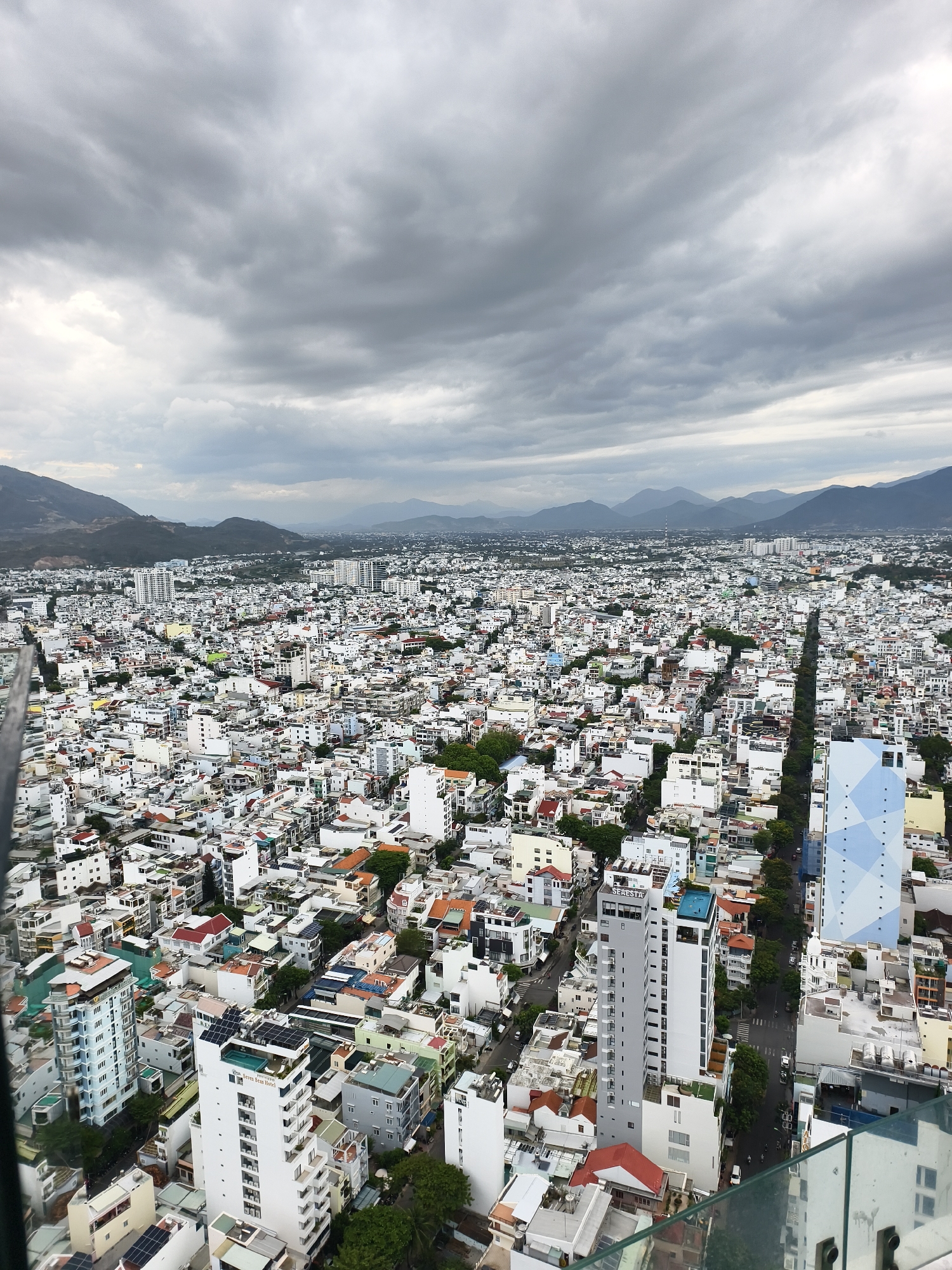 This high-angle, expansive view captures a dense urban cityscape under a heavily overcast, grey sky. The scene is dominated by countless buildings, ranging from closely packed low-rise structures with varying roof colors to numerous taller, modern high-rises. A chain of distant mountains stretches across the horizon, appearing hazy beneath the cloudy sky.
The city displays a complex network of streets and alleys, with a long, tree-lined main road visible cutting through the urban fabric on the right side. Several notable buildings stand out; a tall, light blue building with a distinctive patterned facade is prominent on the right. Closer to the foreground, multiple high-rise buildings feature rooftop swimming pools and solar panels. Visible text includes "Seven Seas Hotel" on a tall white building, and "SERESEY" (likely part of a hotel name) on an adjacent grey building, both with rooftop pools.
The image is taken from a high vantage point, possibly an observation deck or a tall building, suggested by the glass railing in the foreground. The weather is overcast, with diffused daylight, implying a grey or potentially rainy day. No specific human activities are discernible at this scale. The setting is a bustling, modern city, consistent with the coastal urban environment of Nha Trang, Vietnam.