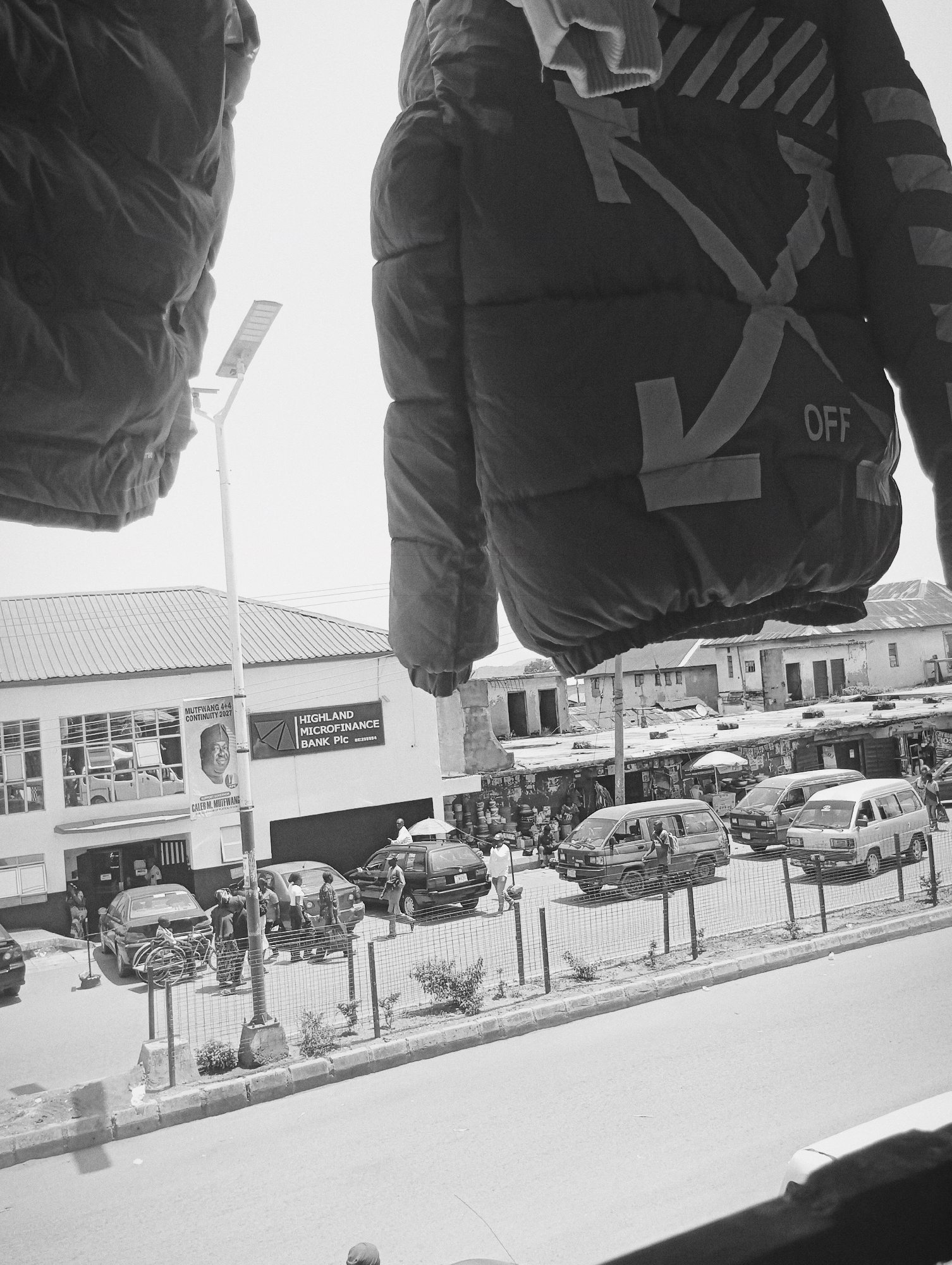 In Bukuru, Nigeria, on a clear day, a street scene unfolds. The foreground is dominated by the lower portion of two dark, puffy jackets hanging, one on the left and one on the right, obscuring the sky. Between them, a tall, slender street lamp with a rectangular light fixture is visible against the bright sky.
Below the hanging jackets, a street is lined with buildings and parked vehicles. On the left, a white building with a prominent sign reading "HIGHLAND MICROFINANCE BANK PIC" is visible, with a banner featuring a portrait of a man and the text "MUTFWANG 4+6 CONTINUITY 2027" and "CALED M. MUTFWANG". Several cars are parked along the street in front of this building and other businesses. A group of people are gathered near the entrance of the bank and by the cars.
Further down the street, a row of smaller buildings and market stalls are visible. Several vans and cars are moving or parked on the road. One van has a person standing in its open doorway. Another van, white in color, is parked on the right side of the road. A black sedan with people standing near it is also parked on the right. A metal fence runs along the side of the road, separating the sidewalk from the street. Several small bushes are planted along the edge of the sidewalk. The overall atmosphere suggests a bustling urban environment with commercial activity.