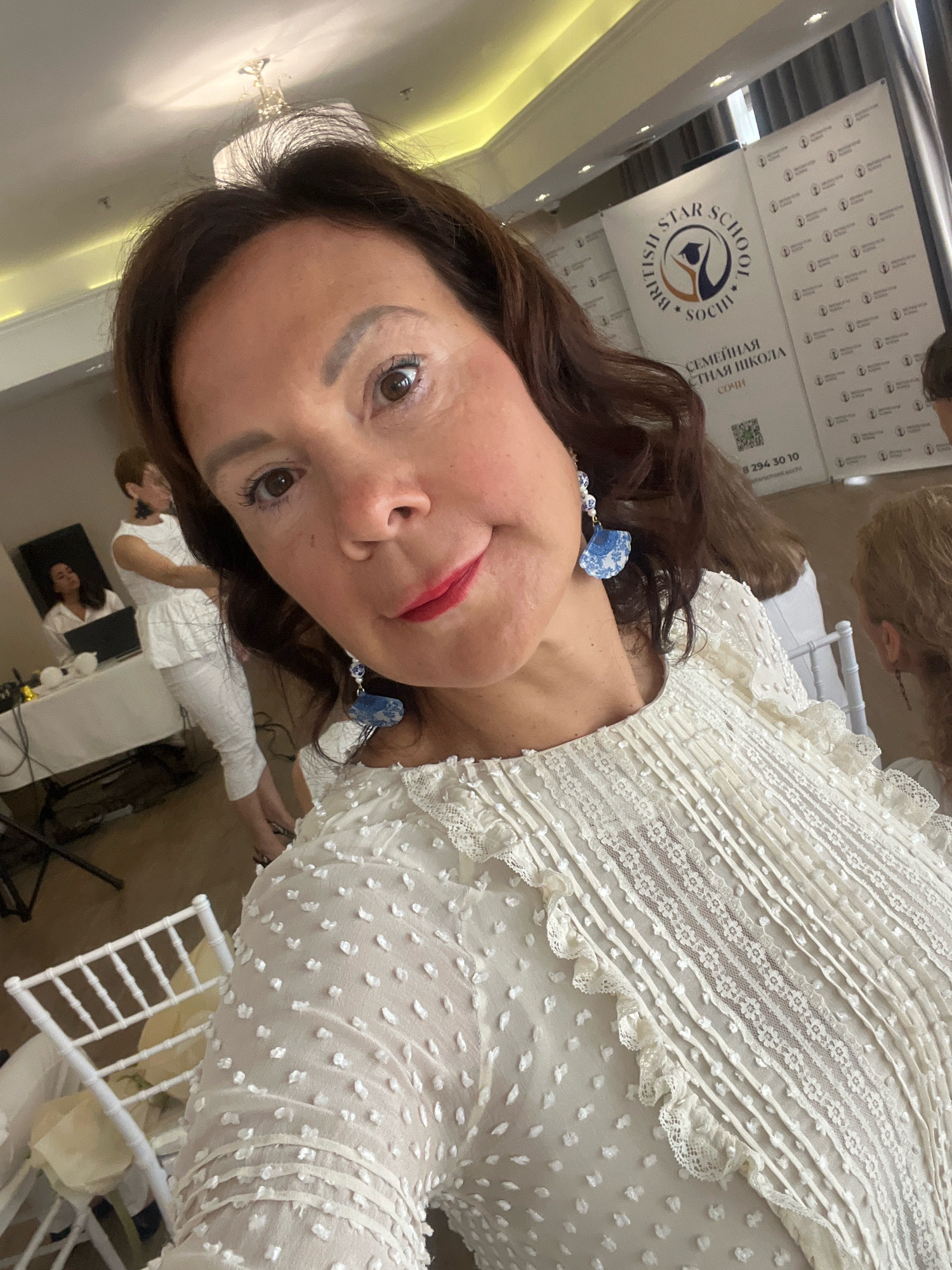 A woman with brown hair and makeup is taking a selfie in a white lace dress. In the background, a banner reads "BRITISH STAR SCHOOL SOCHI" in English and Russian. Other people are visible in the background, including two women in white attire near a table with a laptop and a woman with blonde hair sitting in a white chair. White chairs are also visible in the foreground. The setting appears to be an indoor event space in Sochi, Russia.