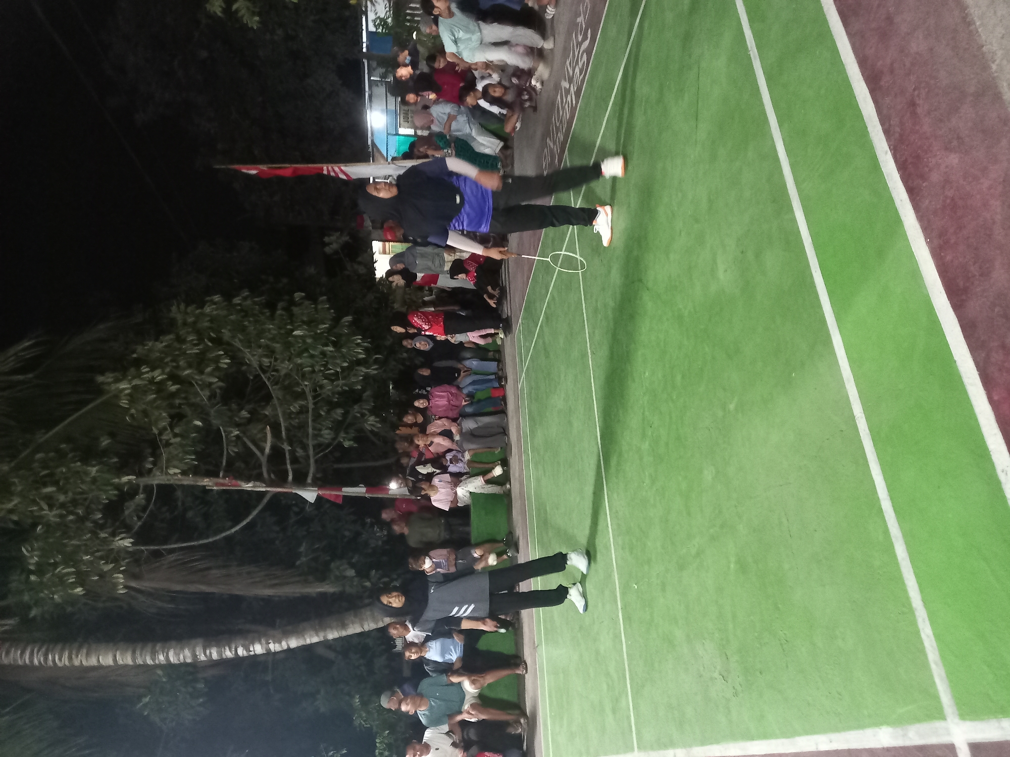 In Banyumulek, Indonesia, a badminton match is taking place at night on a green court marked with white lines. Two female athletes are actively playing. One, wearing a black hijab and a blue and black athletic outfit, stands in the right foreground, holding a badminton racket. The other, wearing a dark hijab and a grey and black tracksuit, is in the left midground, facing towards the right.
A crowd of spectators, composed of men, women, and children, is seated and standing on the left and right sides of the court, observing the game. They are dressed in casual attire. Behind the spectators, there are trees and a palm tree, with a flag pole displaying a red and white flag in the background. The lighting suggests artificial illumination for the night event. The scene appears to be an outdoor community gathering or sporting event.