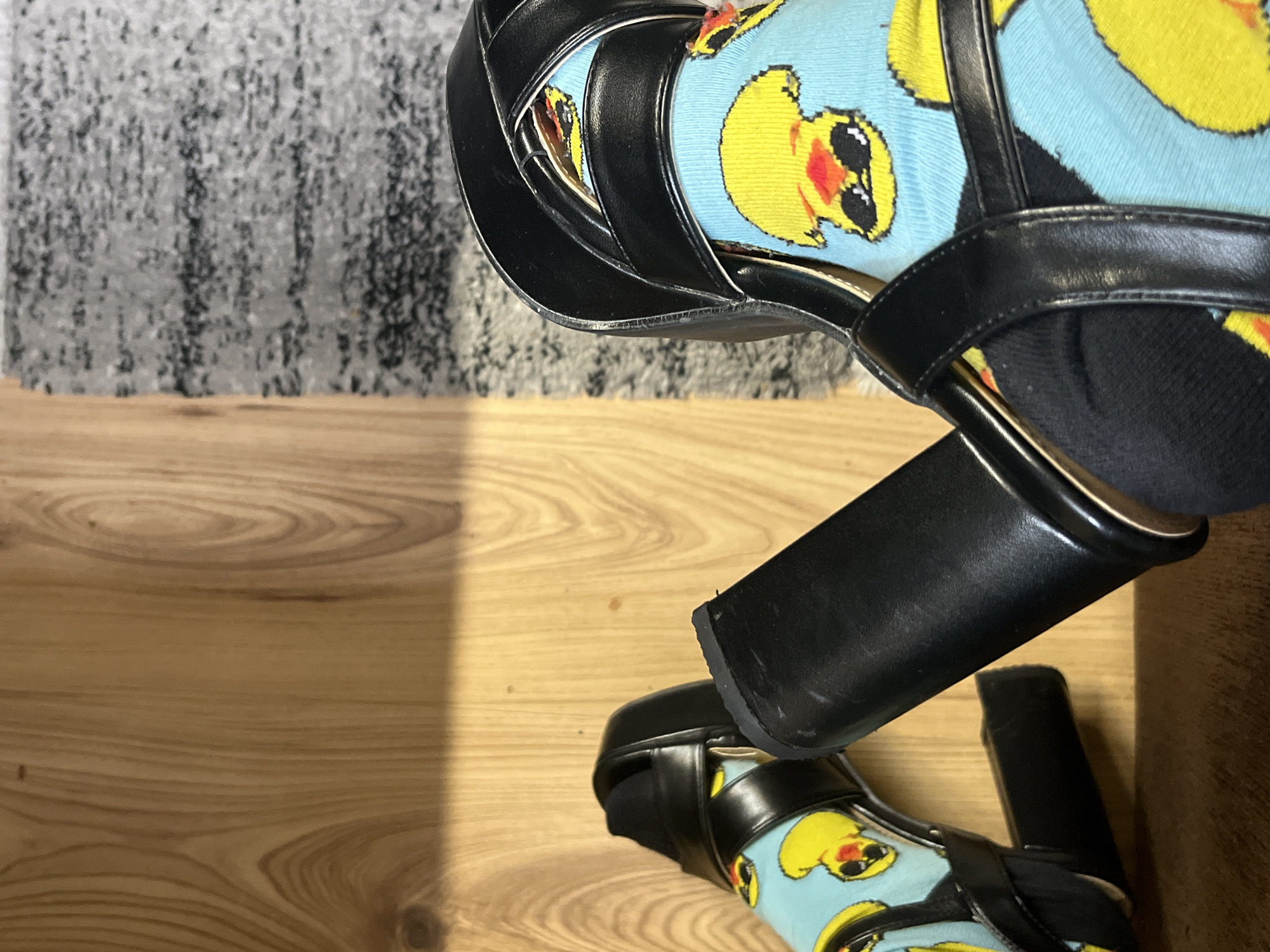 The image depicts two black, high-heeled sandals worn with blue socks patterned with yellow ducklings. The shoes and socks are positioned on a light-colored wooden surface. A gray rug is visible in the upper left corner of the frame. The location is not discernible based on the image.