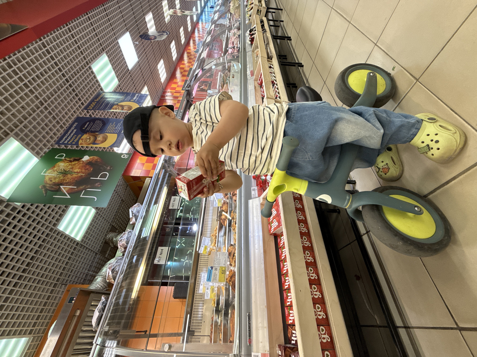 A low-angle, slightly rotated photograph captures a child seated on a tricycle in what appears to be a grocery store aisle, Rusanovo, Russia.
The child, wearing a striped shirt, blue jeans, yellow Crocs, and a black baseball cap, is focused on a small, rectangular carton in their hands.
The tricycle is mostly blue with bright green accents on the handles and wheels. The tires are a dull, worn gray.
The store aisle is visible in the background, with chilled displays of various foods and shelves of packaged goods. Signage in Russian is noticeable above the food displays. The floor is tiled, and overhead lighting is provided by fluorescent fixtures. The shelves beside the child are stacked with small, identical red and white boxes labeled "go."
The scene conveys an everyday moment of childhood curiosity in a commercial setting.