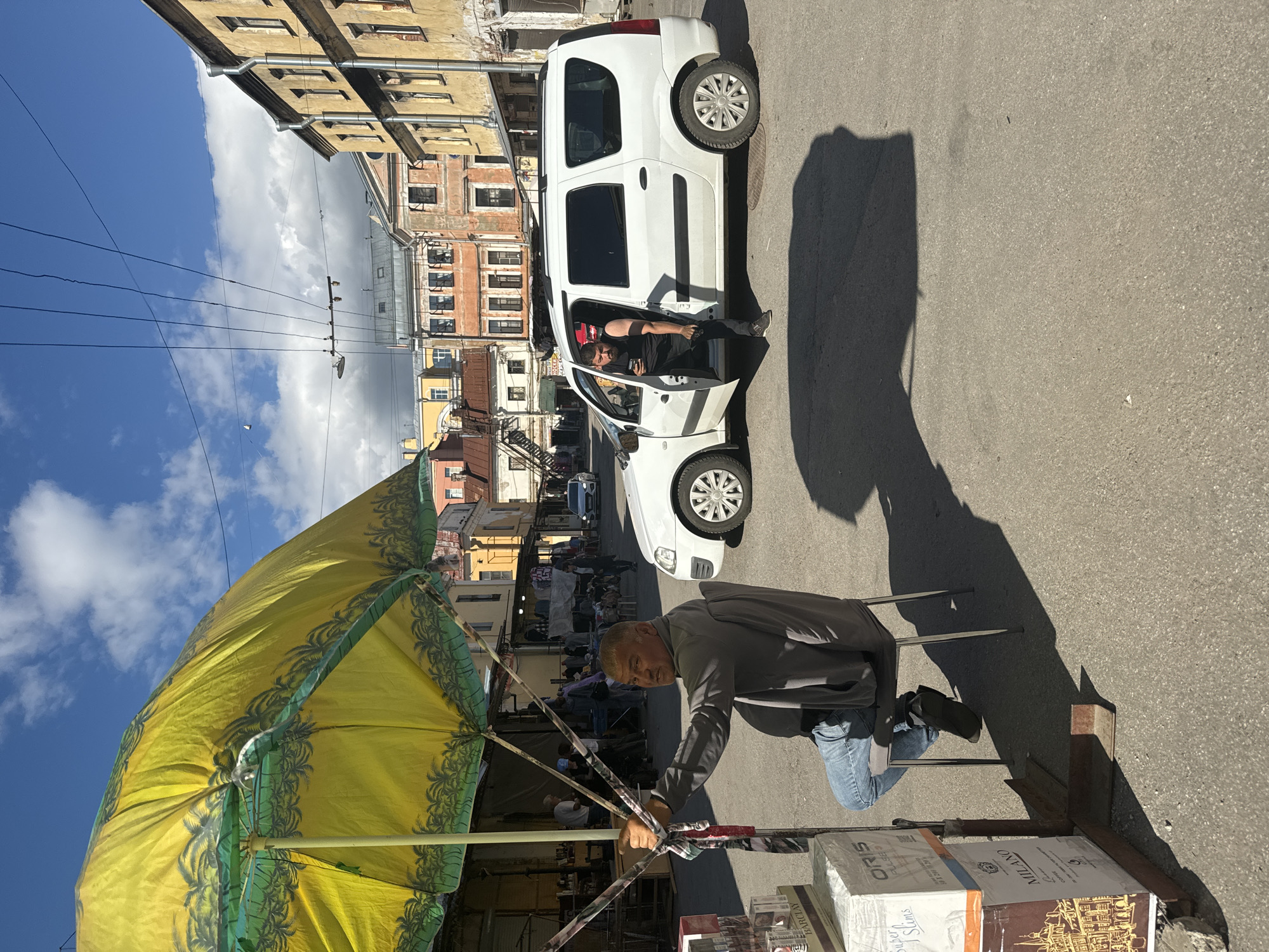 The image depicts a street scene in Saint Petersburg, Russia, likely a market or an area with shops. In the foreground, a man is sitting on a chair under a large, open yellow umbrella with a palm tree pattern. He is wearing a gray jacket and blue jeans. In front of him are stacks of boxes, one of which is labeled "ORIS PULSE" and another "MILANO". To the right, a white van is parked with its driver-side door open. A man in a black t-shirt is leaning out of the open door, holding a cup. The van is parked on an asphalt road. In the background, there are several buildings with multi-story facades, typical of Saint Petersburg's architecture, painted in various colors including yellow and pink. Overhead, power lines and cables are visible against a blue sky with scattered clouds. The overall impression is that of a warm, sunny day in a bustling urban environment.