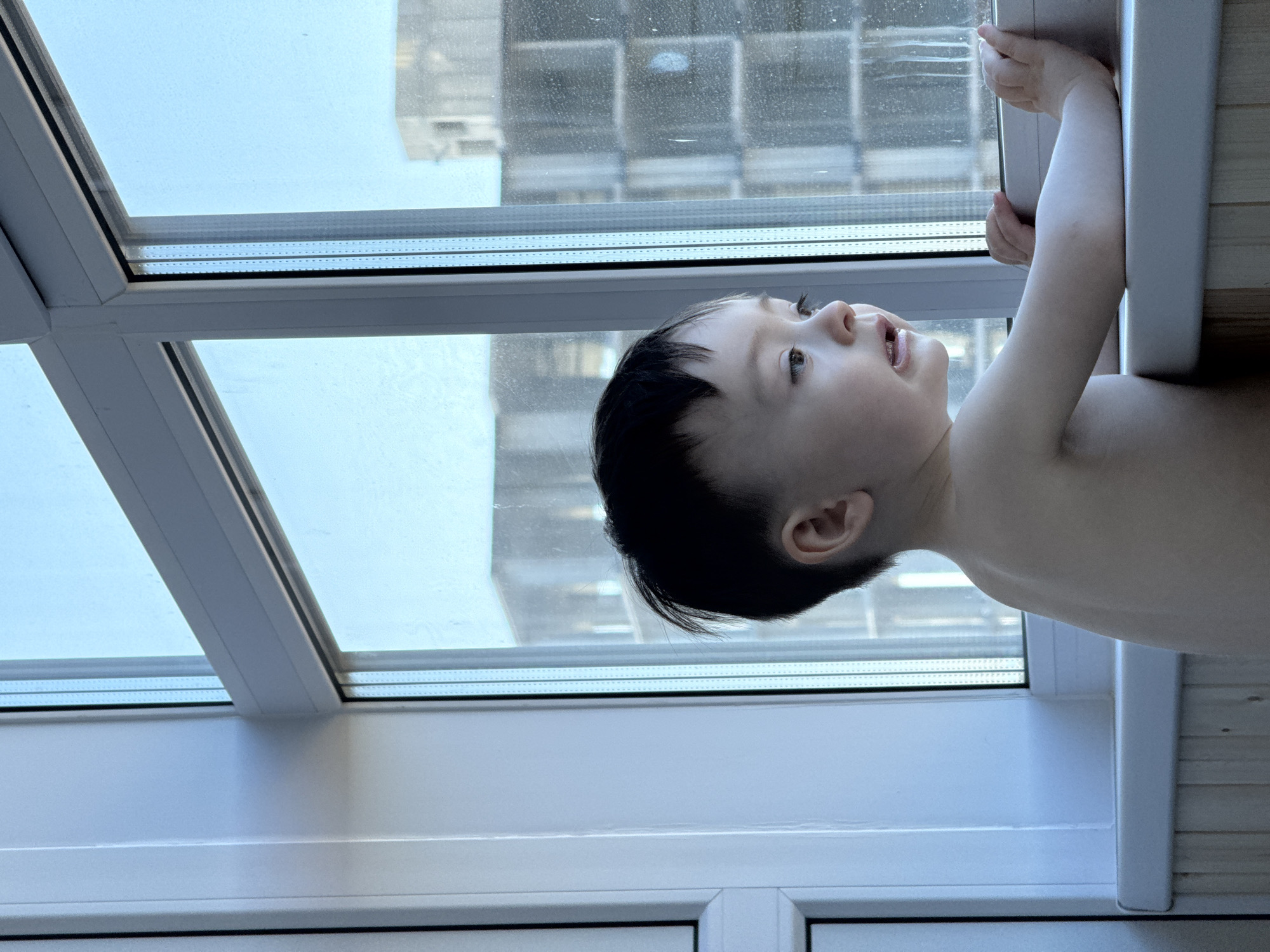 A young child is looking out of a window. The window has a view of a building. The child has dark hair and is wearing no clothes. The setting is Izhevsk, Russia.