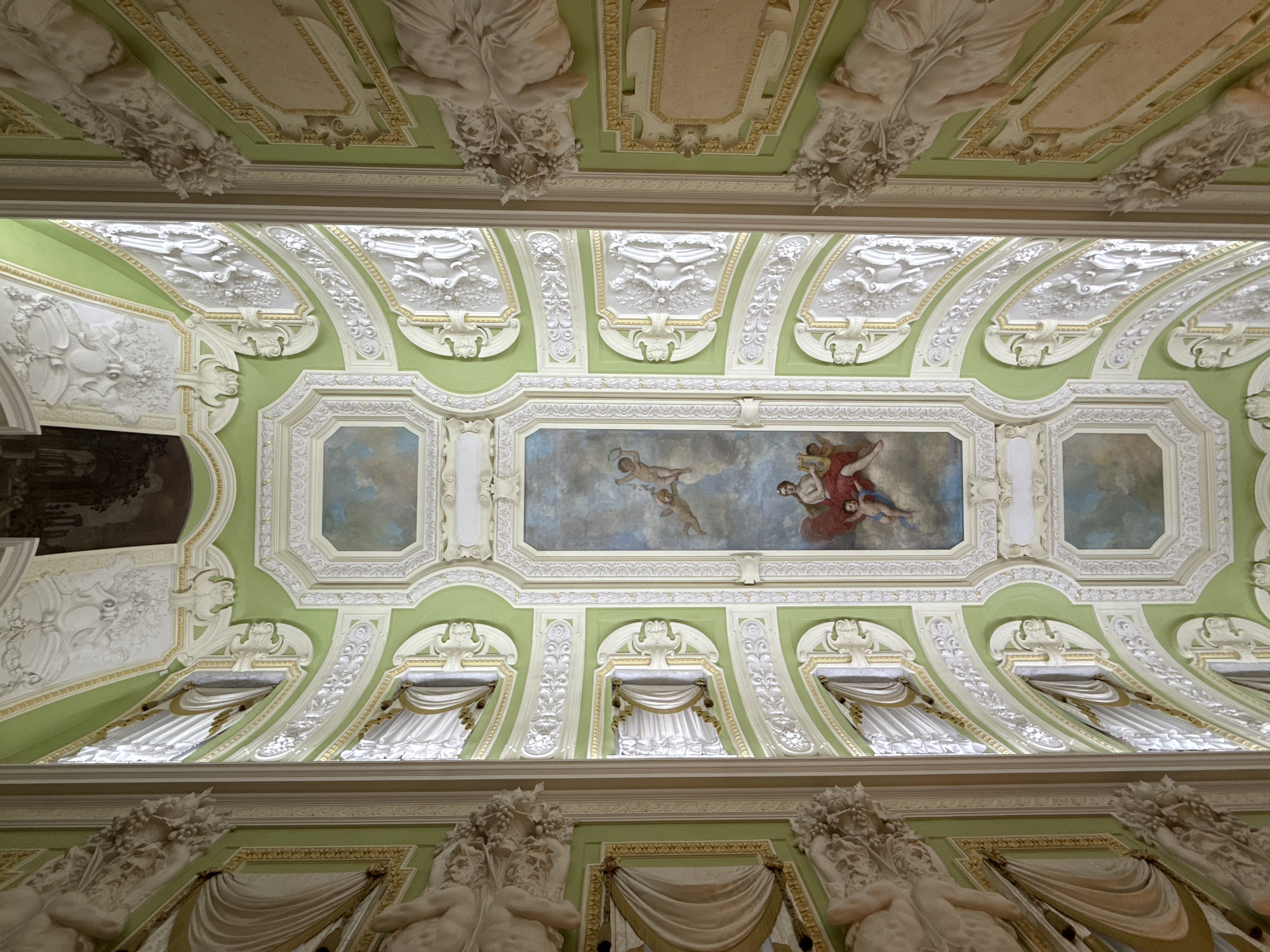 The image shows a highly ornate, ornate ceiling with a fresco in the center. The ceiling is painted in a pale green color and is adorned with intricate white stucco work, gilded accents, and various sculptural elements. The central fresco depicts figures, including what appear to be cherubic figures and a woman in red, set against a cloudy sky. The overall style suggests a Baroque or Rococo influence. The lighting indicates an interior setting, likely a grand hall or ballroom. The image does not contain any identifiable landmarks that would allow for the determination of its location in Nizhny Novgorod, Russia.