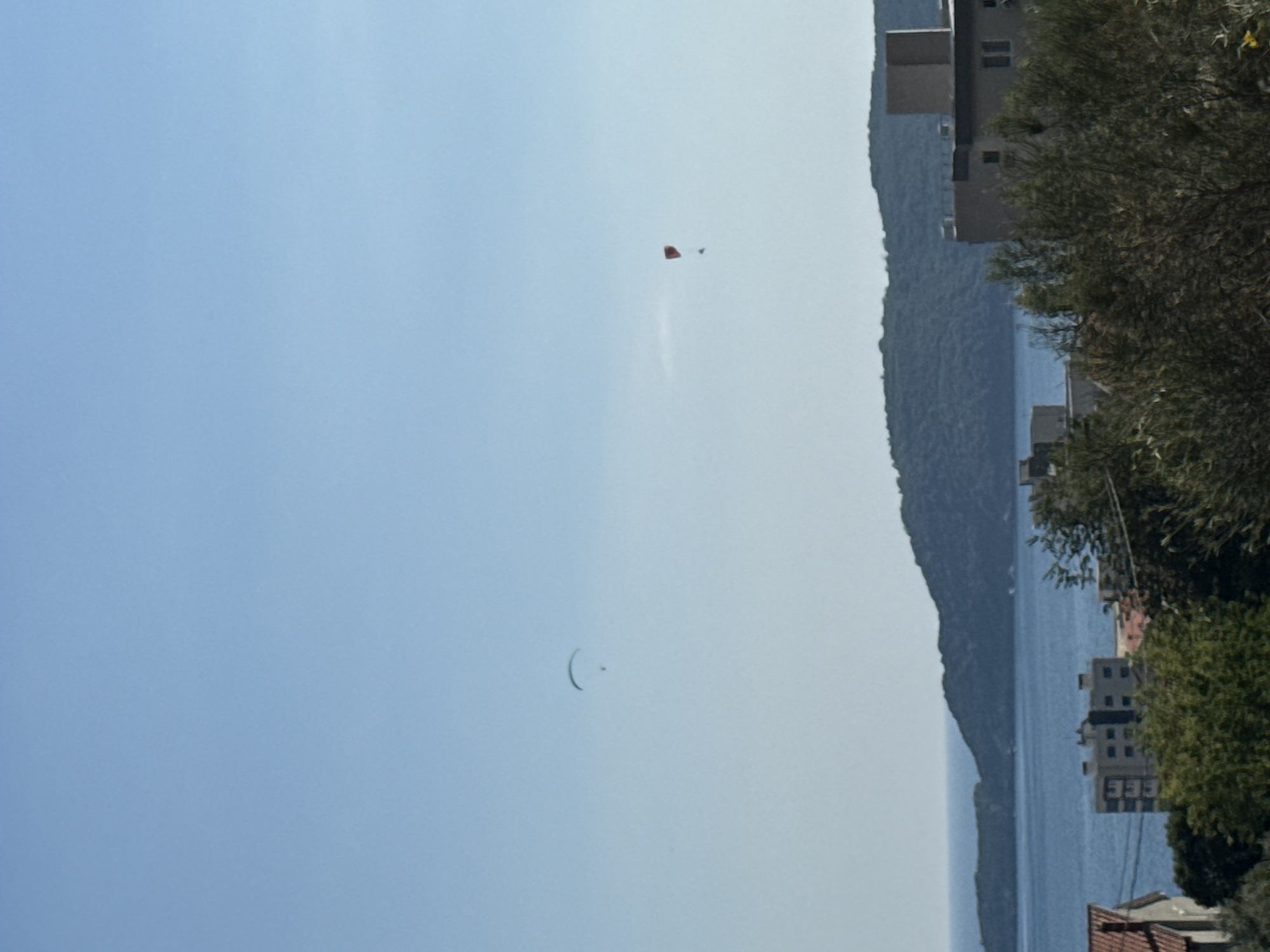 An outdoor, eye-level, wide shot captures a clear, bright sky over a coastal landscape. In the upper left of the frame, a paraglider with a curved wing and a suspended figure is visible. Further to the right, a second paraglider, this one red with a yellow accent, is also in the air, with a figure beneath it. The middle ground is dominated by a calm, blue body of water and a dark, forested hill. In the foreground, buildings with light-colored facades are nestled amongst lush green trees, with the roof of a building with red tiles visible on the far left. The scene appears to be the town of Rafailovići, Montenegro.