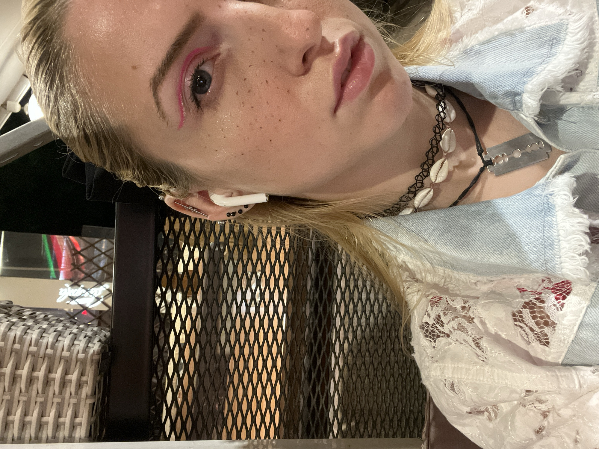 A close-up photograph features a fair-skinned woman with blonde hair, exhibiting makeup consisting of pink eyeliner and lipstick. She is wearing a light blue denim jacket trimmed with white lace and two necklaces: a black choker adorned with white shells and another with a razor blade pendant. An earbud is visible in her ear. The background includes a textured metal grill and a glimpse of a wicker chair. The setting is plausibly in Plovdiv, Bulgaria, based on visual elements, though without additional data, precise location cannot be determined.