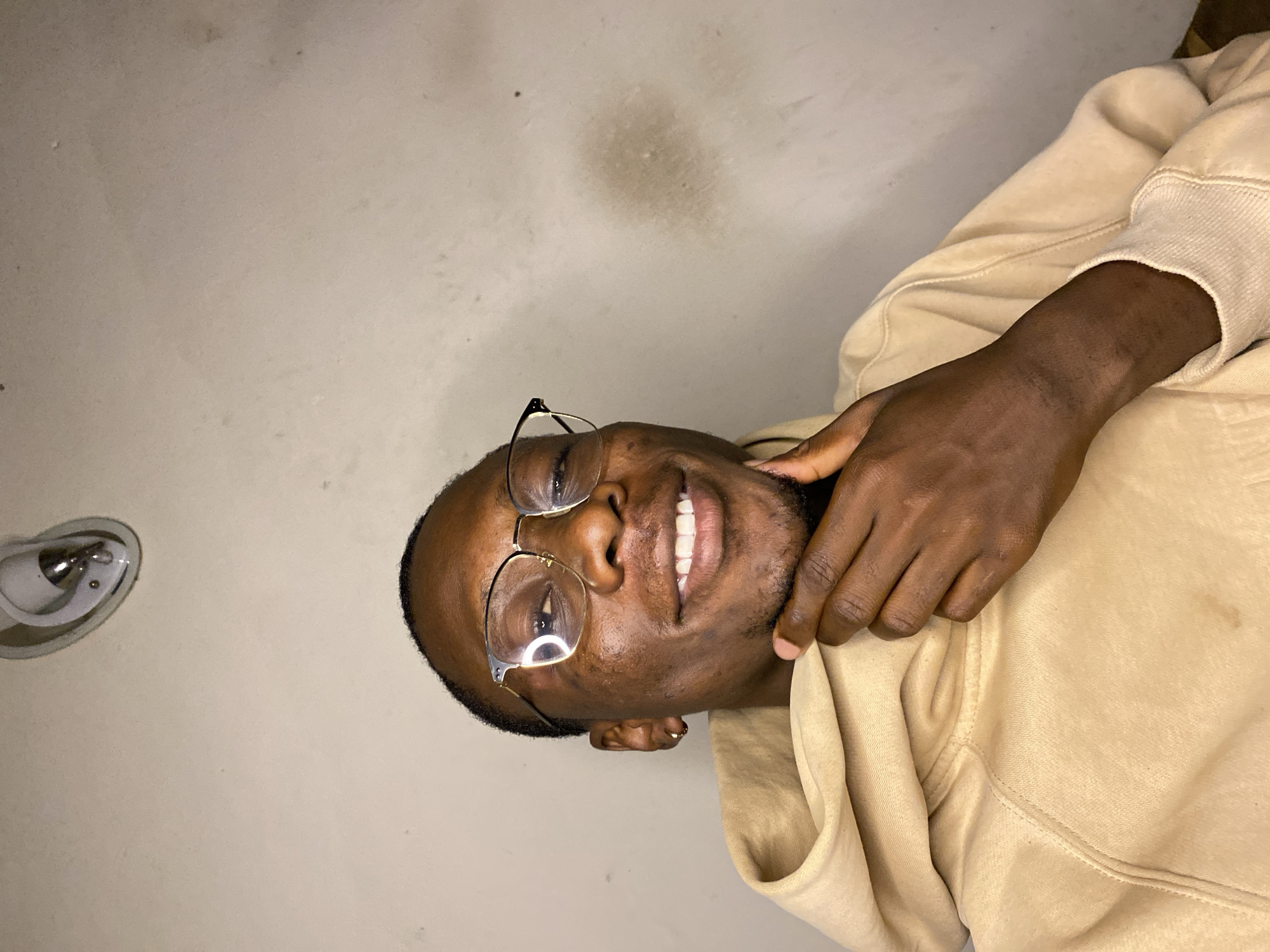 The image, taken in Bukuru, Nigeria, captures a smiling Black man from the chest up, positioned at an oblique angle as if looking upwards towards the camera. He wears a light beige or tan hooded sweatshirt. His face, illuminated by artificial light, shows a broad, cheerful smile, revealing his white teeth. He has short hair and is wearing clear-framed glasses with metallic details; a light reflection is visible on one lens. His right hand is gently placed on his jawline or neck.
The background is a plain, light-colored wall that shows some subtle texture, along with a few smudges and darker stains, suggesting an aged or well-used indoor space. On the far left of the background, a small, silver-toned fixture, possibly a light or smoke detector, is partially visible. The overall mood is relaxed and happy, indicated by the man's smiling expression. There is no visible text in the image.
