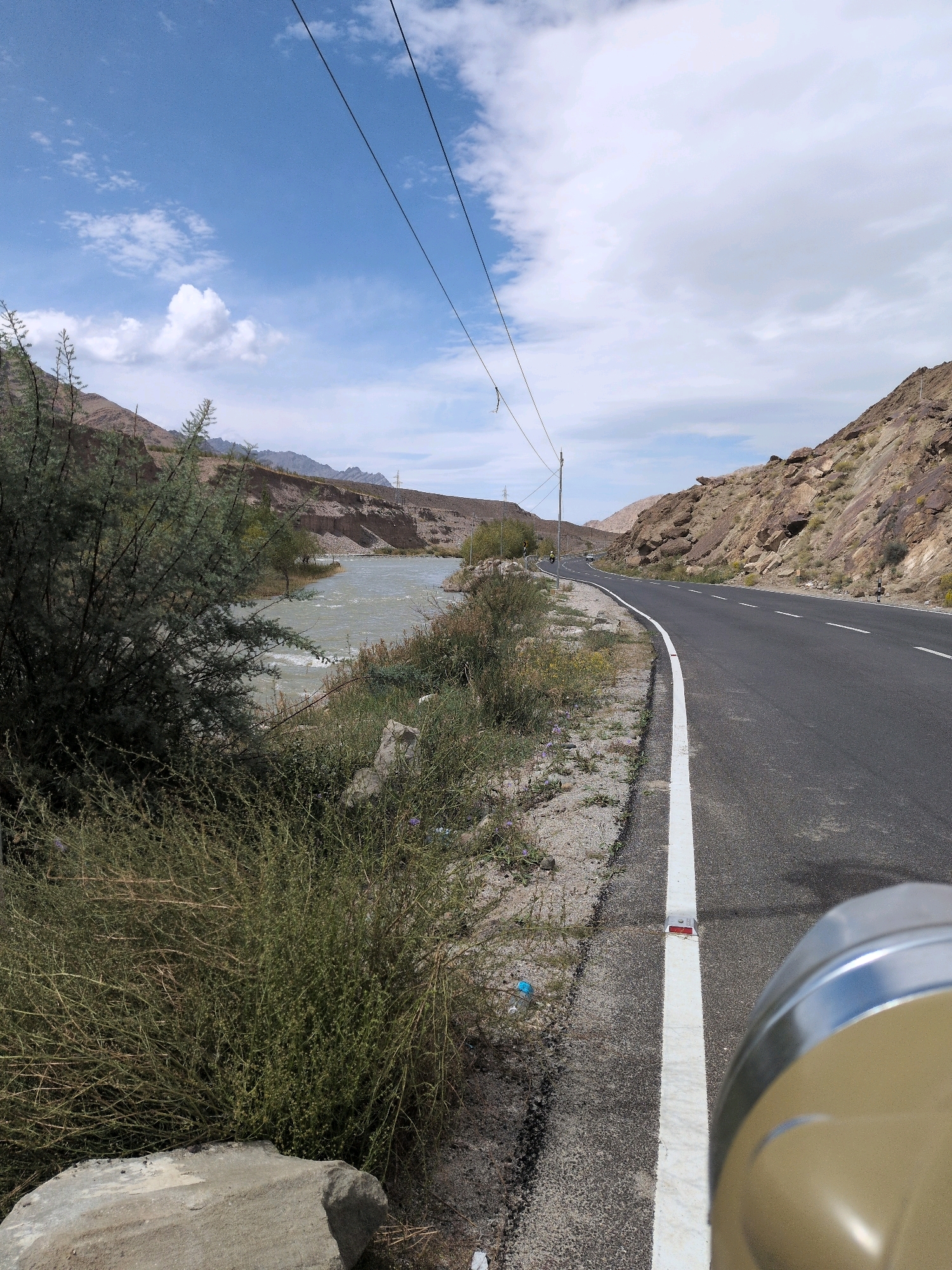 A paved road with a white line marking the edge curves to the right, bordered by a rocky, barren mountainside on the right and sparse vegetation, a river, and more mountains in the distance on the left. Power lines are strung across the sky. A blurry, metallic object, possibly part of a vehicle, occupies the bottom right corner of the frame. The scene appears to be a rural roadside in Upshi, India, with a few vehicles visible on the road in the distance. The sky is partly cloudy, with blue sky and white clouds.