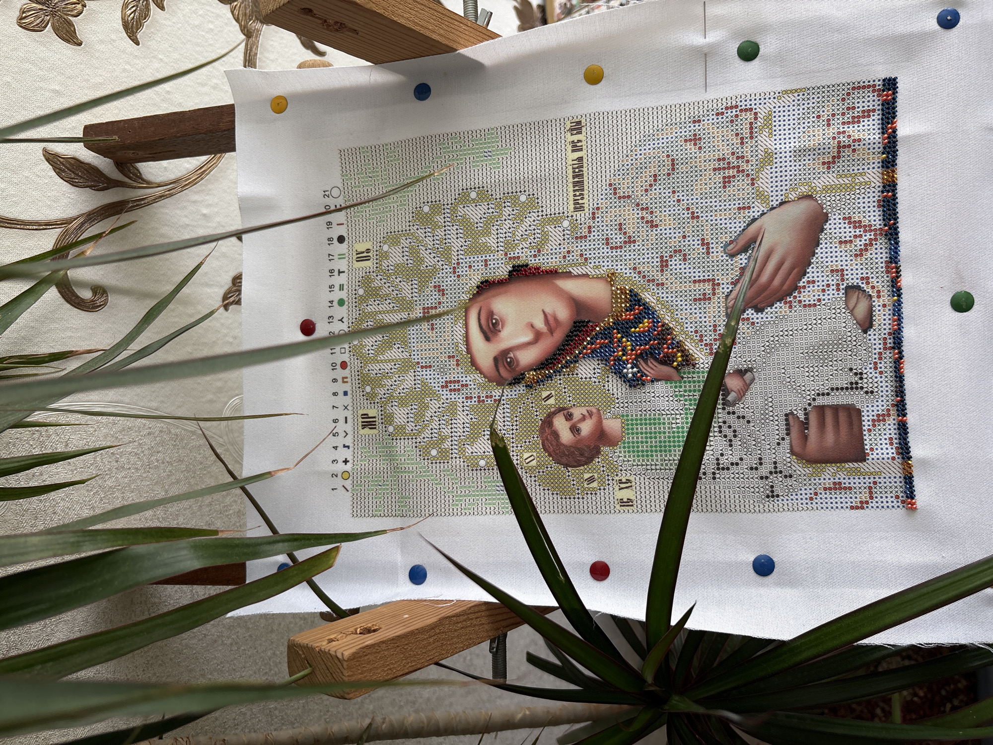 A cross-stitch project depicting religious figures is displayed on a wooden frame, pinned to a white cloth. The partial figures include a woman and a child. Text in what appears to be Cyrillic script is located in the top-right portion of the cross-stitch. The stitching is in progress, showing completed areas with colored threads and unfilled areas represented by symbols and grids. The frame holding the cloth is resting on a surface, possibly a table, and a green leafy plant occupies the foreground, partially obscuring the project. Pins of various colors – red, blue, yellow, and green – are securing the cloth to the frame. No information related to Chaikovsky, Russia, is included in the description, as the provided image does not contain location-specific details.
