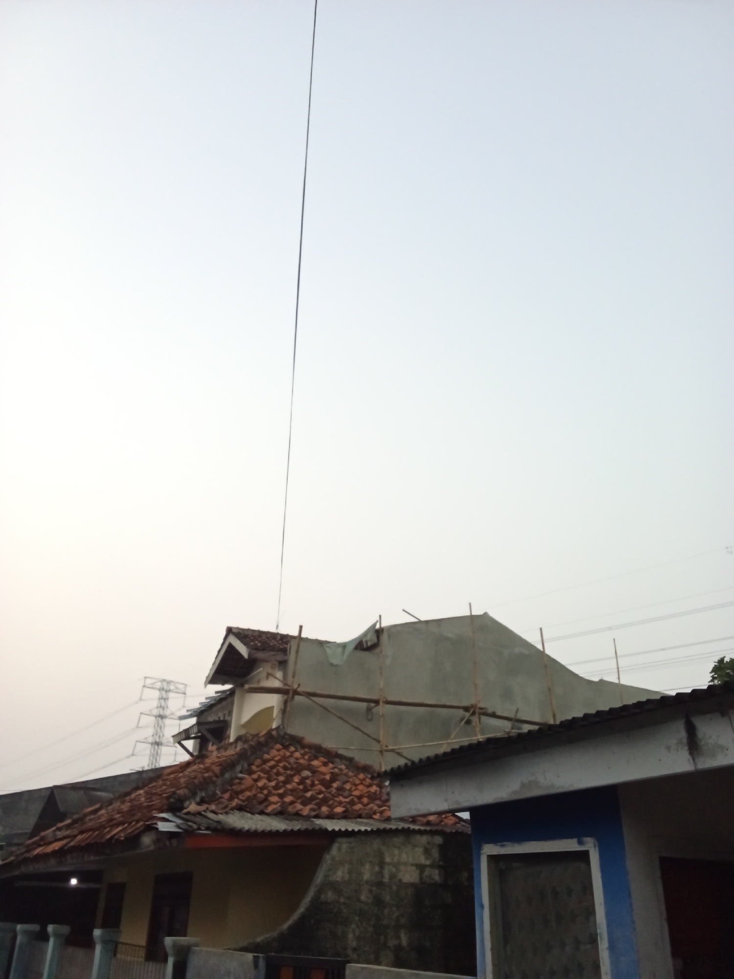 This is a medium-angle, eye-level shot of a residential area in Depok, Indonesia, under an overcast sky. The foreground features the roof and upper facade of a house with a blue-trimmed window opening. Behind it, the roofs of several other houses are visible, some covered with terracotta tiles and others with corrugated metal. Construction scaffolding made of bamboo poles is attached to one of the buildings, suggesting ongoing renovation or building work. In the middle ground, a tall electrical tower and transmission lines stretch across the horizon, hinting at the presence of significant infrastructure. A thin black wire stretches diagonally from the top of the frame down towards the buildings. The overall impression is one of urban residential development with industrial elements present in the background. There are no people visible in the frame.