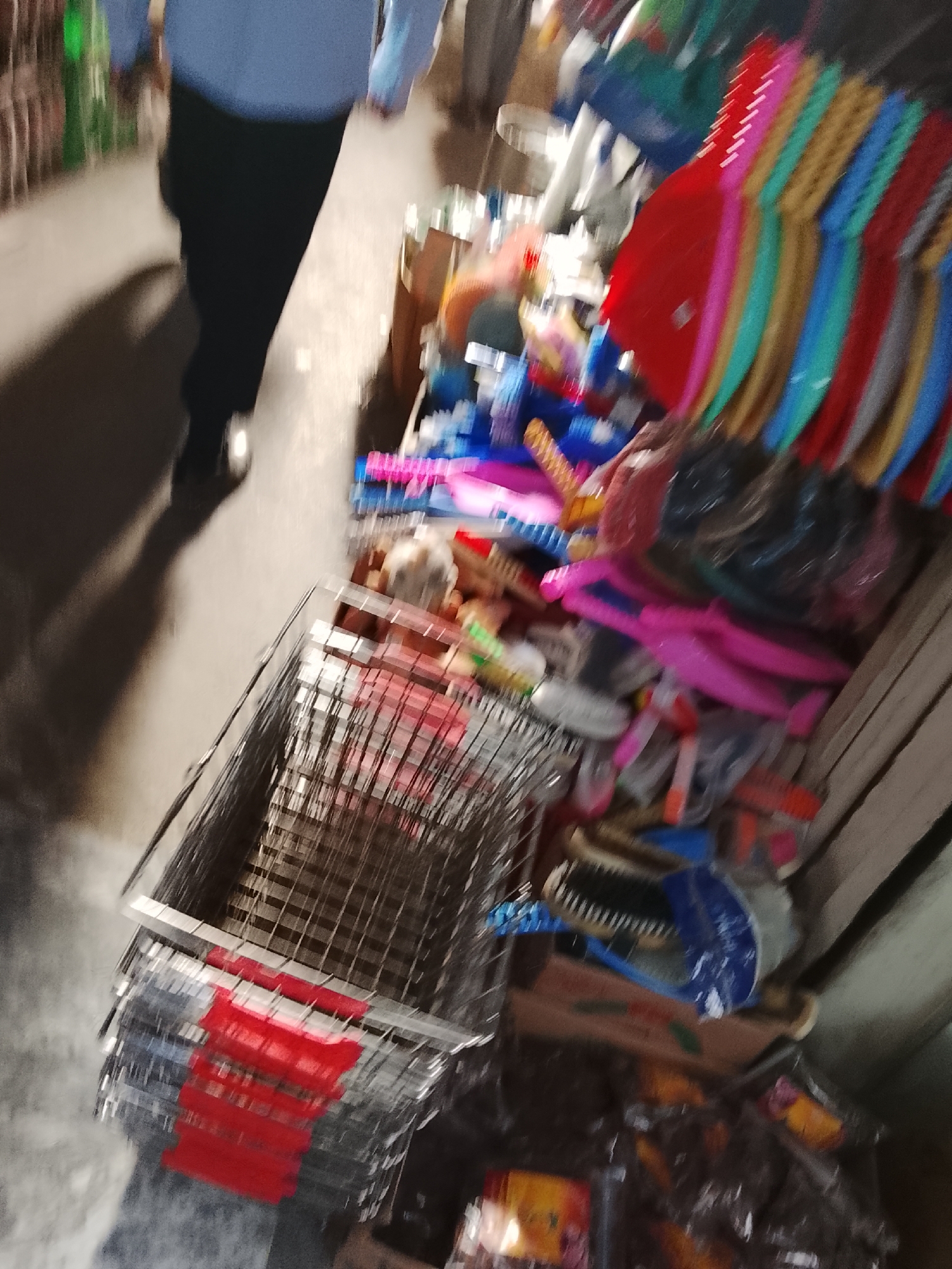 The image shows a shopping cart in the Gulgasht Colony, Pakistan. The cart is made of metal and has red plastic pieces on the front. It appears to be filled with various household items, including colorful dustpans, plastic watering cans, brushes, and cleaning supplies. A person wearing a light-colored shirt and dark pants is walking past the cart. The scene seems to be taking place in a shop or market area, as there are other items and displays visible in the background.