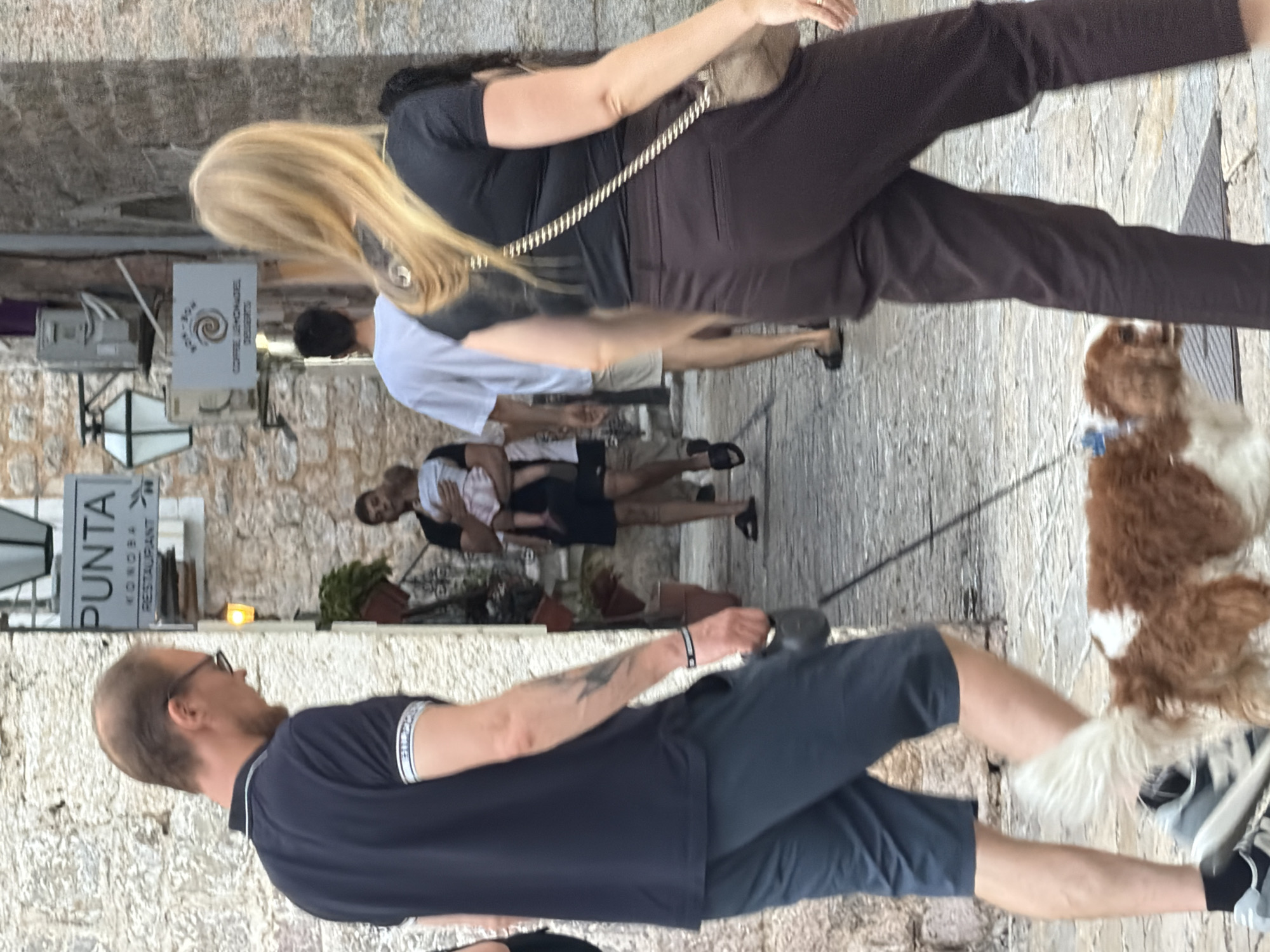The image captures a street scene in Budva, Montenegro. In the foreground, a man wearing a dark blue polo shirt and dark blue shorts walks a small, brown and white dog on a leash. The dog's tail is held high, and it appears to be trotting alongside the man. The man's left arm is visible, showing a tattoo. Behind him, a woman with blonde hair, wearing a black top and dark brown pants, walks away from the camera, carrying a light-colored crossbody bag.
Further in the background, a man in a white t-shirt and light-colored shorts walks away. To his left, another man, wearing a dark t-shirt and dark shorts, is holding a baby. The baby is dressed in light pink. The environment suggests a narrow street with stone buildings. A sign with "PUNTA KONOBRA RESTAURANT" is visible on the left side of the street. Another sign with a spiral logo and text that appears to read "BON-BON COFFEE LEMONADES DESSERTS" is present further down the street on the right. The ground is paved with cobblestones. The overall lighting suggests it is daytime, possibly late afternoon or early evening, with some shadows cast on the ground.