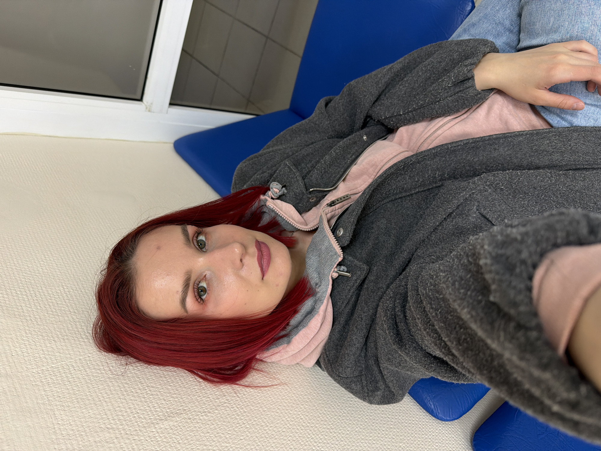 A young woman with bright red hair and light green eyes is pictured lying down indoors in Chaikovsky, Russia. She looks directly at the camera with a neutral to slightly expressive gaze, possibly taking a selfie or being photographed from an overhead perspective. Her makeup includes defined eyebrows, winged eyeliner, and pink lipstick.
She is dressed in layers, wearing a dark grey, textured, long-sleeved coat or jacket with a hood, unzipped to reveal a pink hooded top underneath. Her lower body is partially visible, showing light blue jeans. Her left hand is resting near her hip.
The woman is positioned on a light-colored, textured surface, possibly a floor or a wide bench, with her head resting on it. Her body extends onto a pair of bright blue, padded seats or chairs, which appear to be typical of a waiting area or public space. In the background, to her upper left, is a white-framed window, offering a view of an indistinct, possibly gray, exterior. The lighting suggests an indoor setting with ambient light filtering in. There is no visible text in the image.