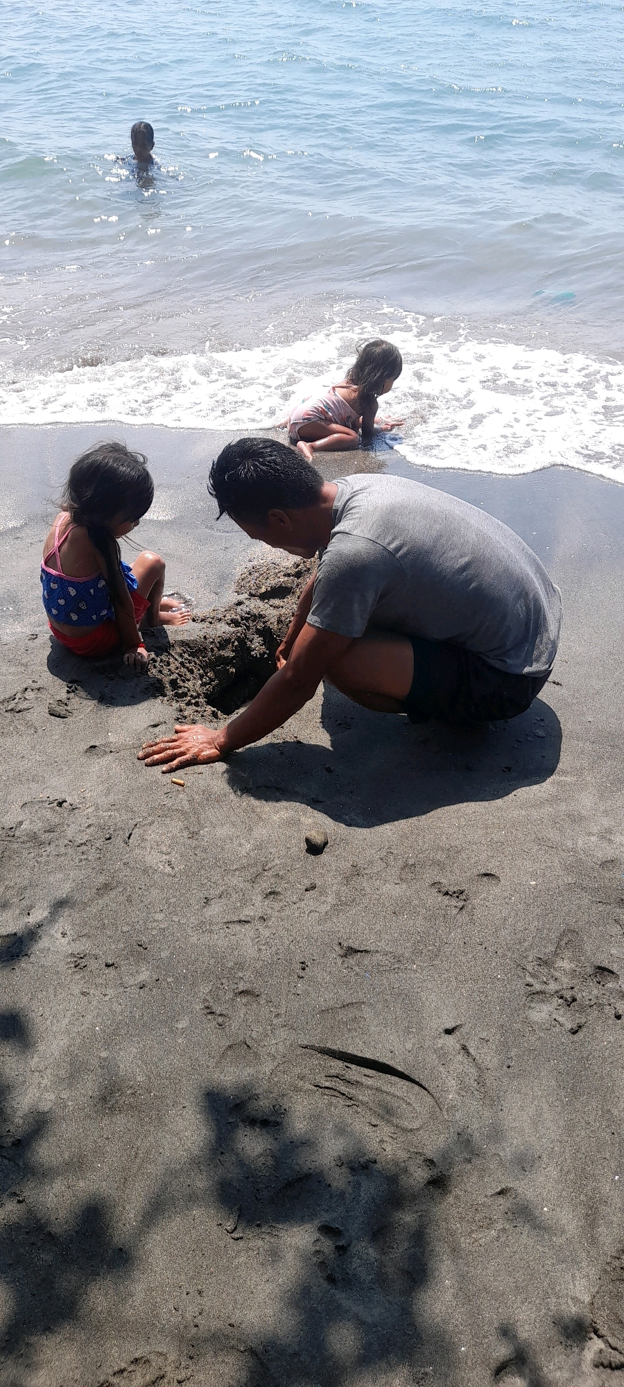 The image depicts a family at Pasir Putih beach in Indonesia. A man is crouching on the dark sand, actively digging a hole with his hands. Beside him, a young girl in a blue and pink swimsuit sits on the sand, also looking at the hole. Further back, another young child, wearing a pink striped outfit, is kneeling near the water's edge, where gentle waves are breaking onto the shore. In the distance, a person is visible in the clear blue water. The environment is a beach with dark sand, wet and dry areas, and the ocean.